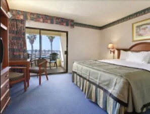 A hotel room with a bed, a desk, a chair, and a view of the outdoors. - Travelodge by Wyndham Ocean Front (Huntington Beach)