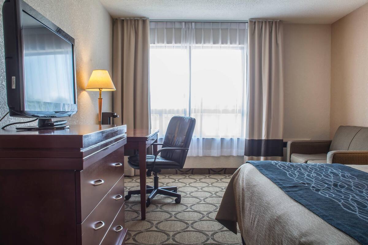 A hotel room with a bed, a desk with a chair, a television, and a window with curtains.