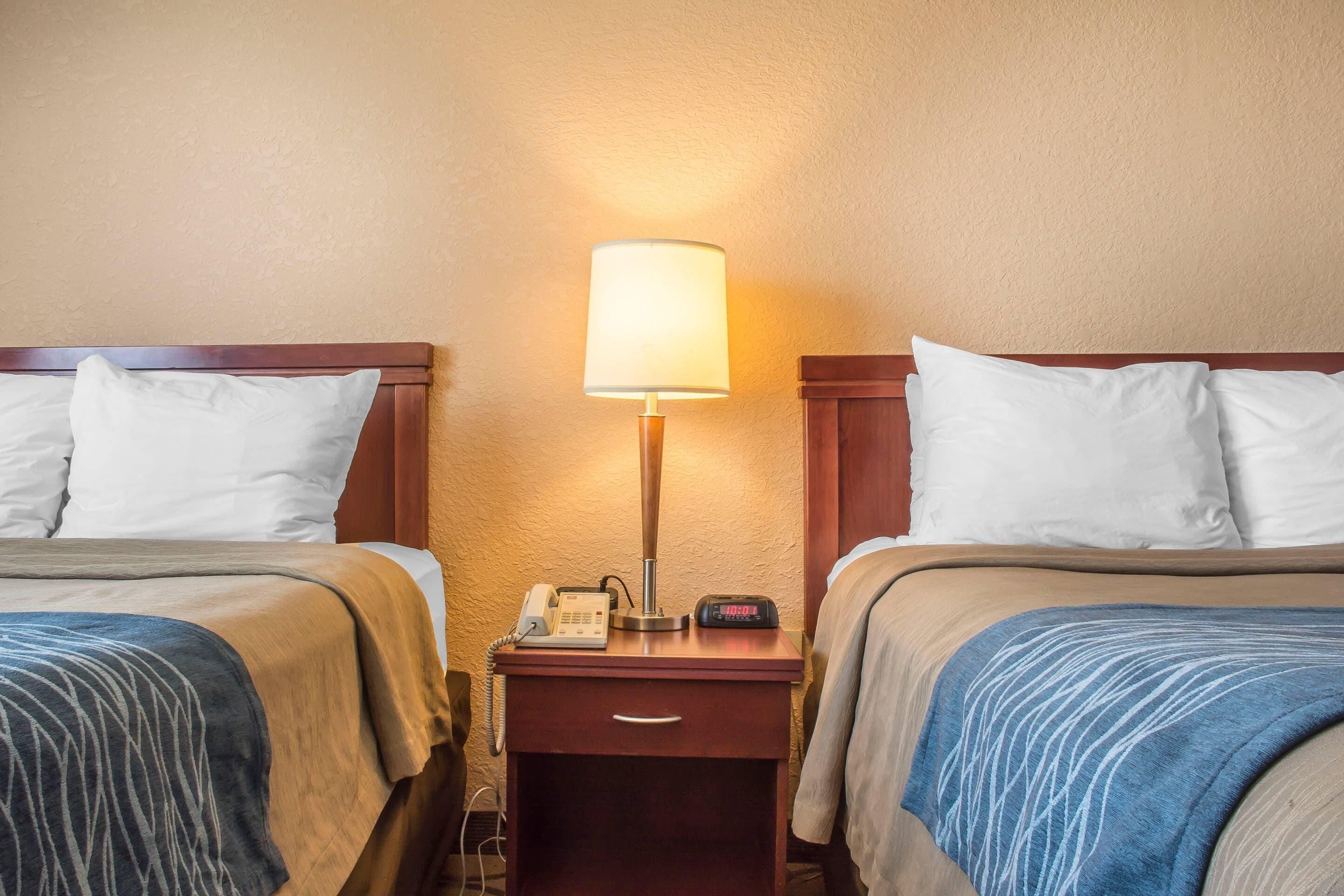 Habitación de hotel con dos camas, una mesita de noche con lámpara, teléfono y reloj digital.