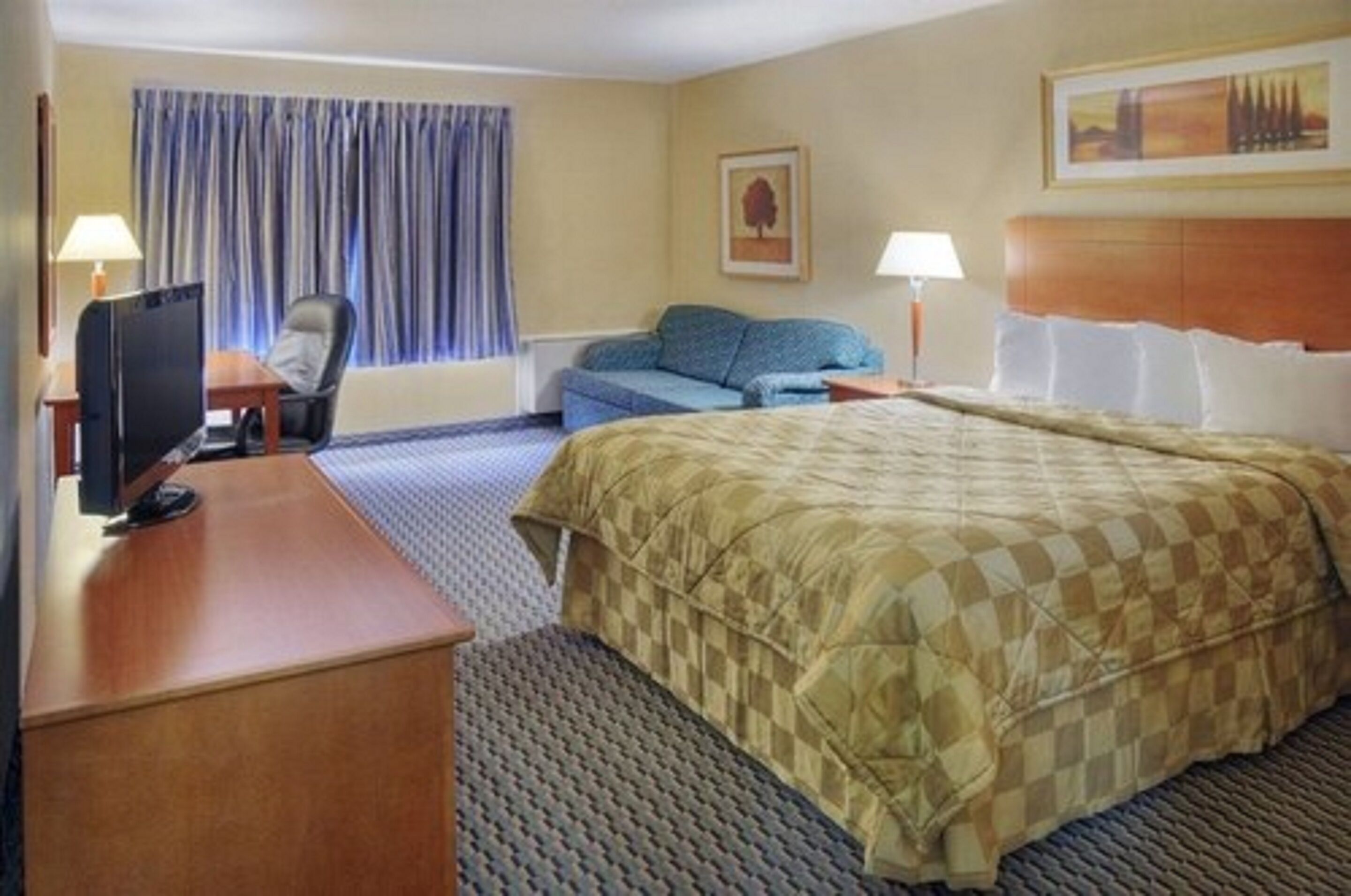 A hotel room with a bed, a desk with a computer, a chair, a sofa, and a window with curtains.
