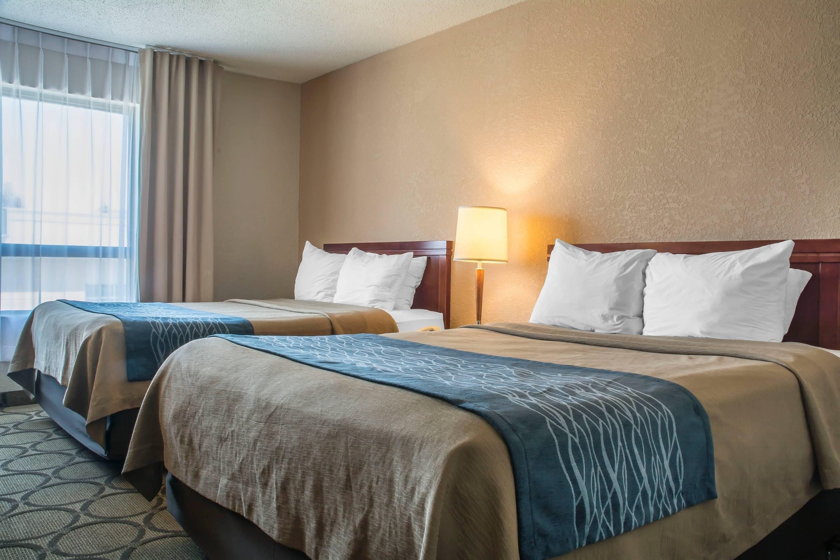 A hotel room with two beds, beige and blue bedspreads, a window with curtains, and a lamp on the nightstand.