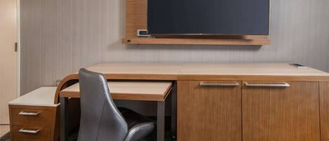 A hotel room with a wooden desk, a black office chair, a flat-screen TV, and a small drawer unit.
