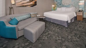 A hotel room with a grey sofa, a bed with white bedding, a small table with two glasses, and a lamp on the nightstand. - Sonesta Select Atlanta Norcross (Norcross)