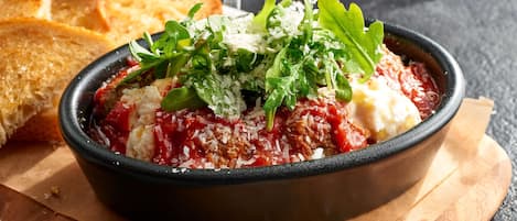 Un plato a base de tomate, adornado con vegetales verdes y queso, acompañado de pan tostado sobre una tabla de madera.
