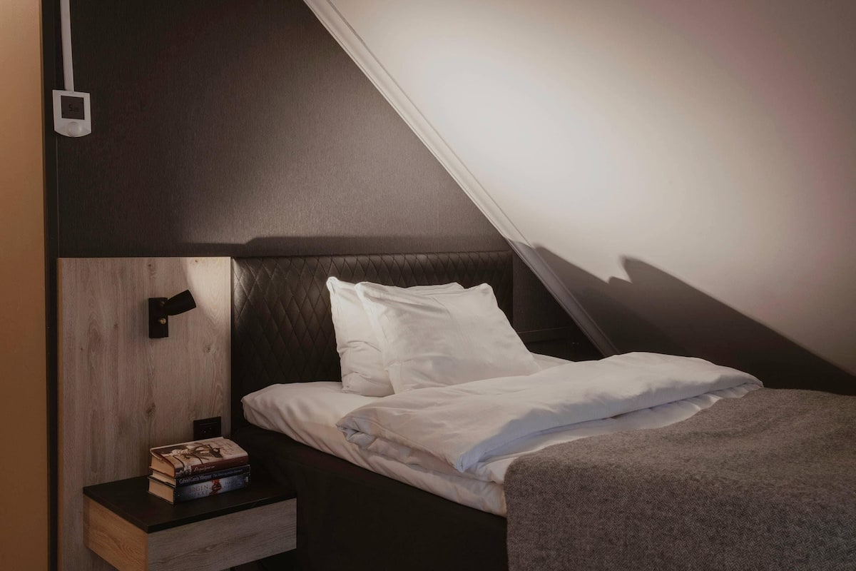 A bedroom with a bed, a nightstand with books, a wall-mounted light, and a dark headboard.