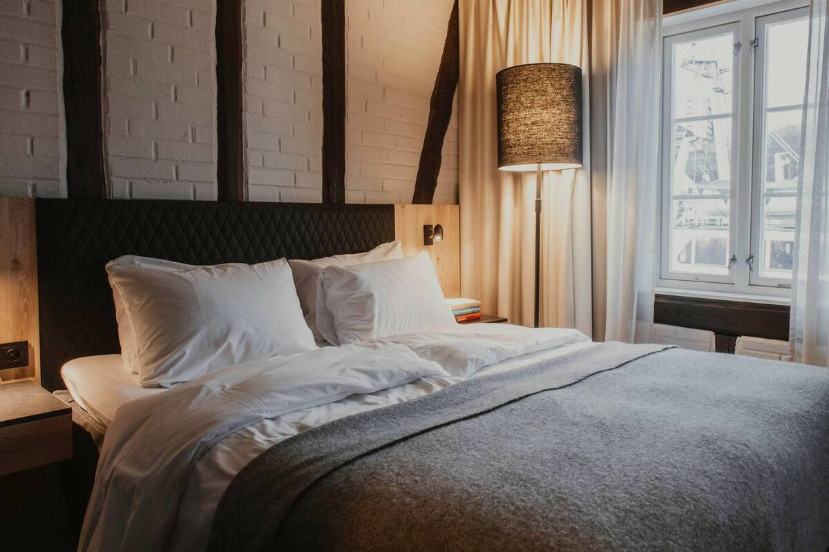 A neatly made bed with white linens, a dark headboard, a bedside lamp, and a window with curtains.
