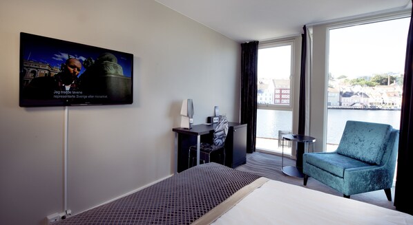 Habitación de hotel con cama, televisión montada en la pared, escritorio con silla, una mesita y vista a una masa de agua a través de amplios ventanales.