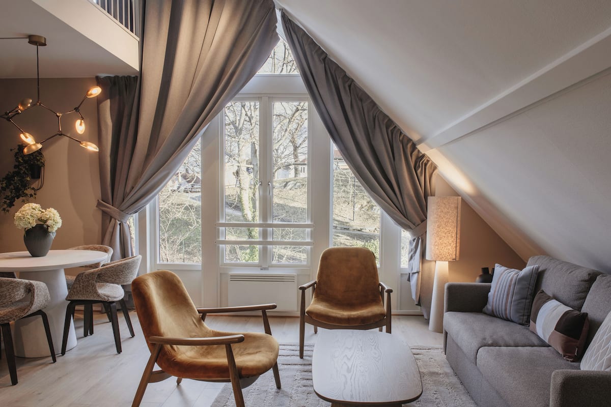 A room with a dining table, two chairs, a sofa, a lamp, and a large window with curtains.