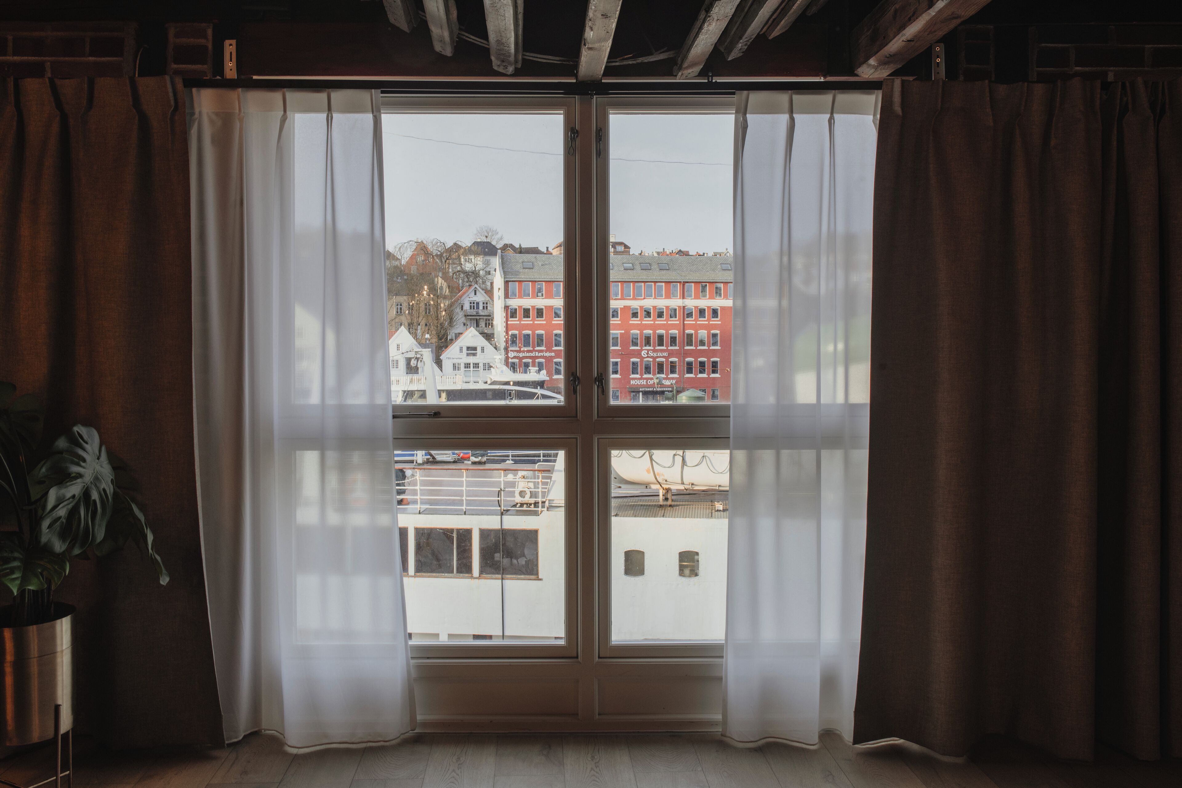 A room with a large window, curtains, a potted plant, and a view of buildings.