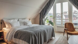 A bedroom with a large bed, a chair, a window with curtains, and a view of buildings. - Home Hotel Skagen Brygge (Stavanger)