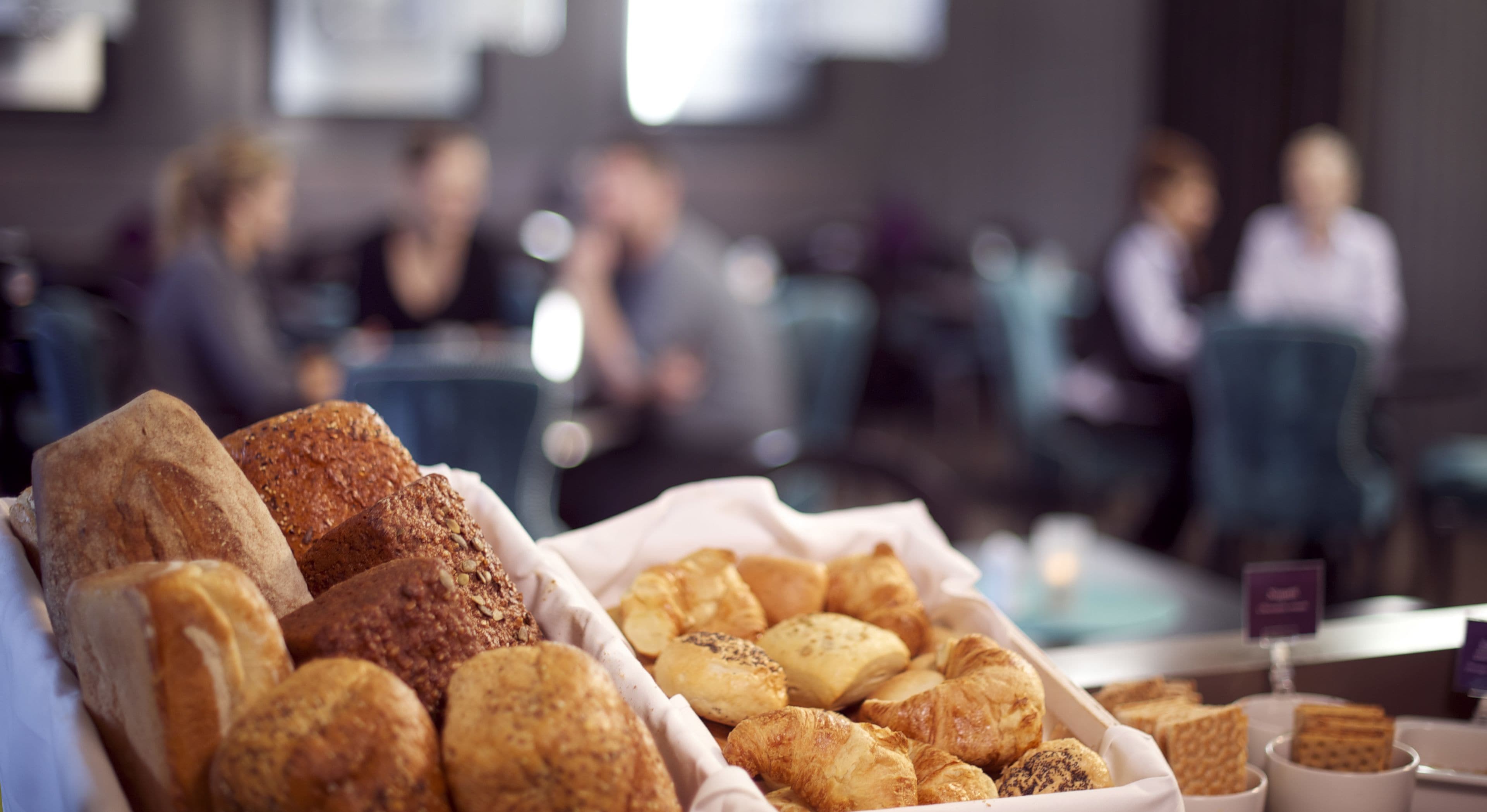 A bakery display with various baked goods in trays, set in a cafe or bakery area with blurred background of people and seating.