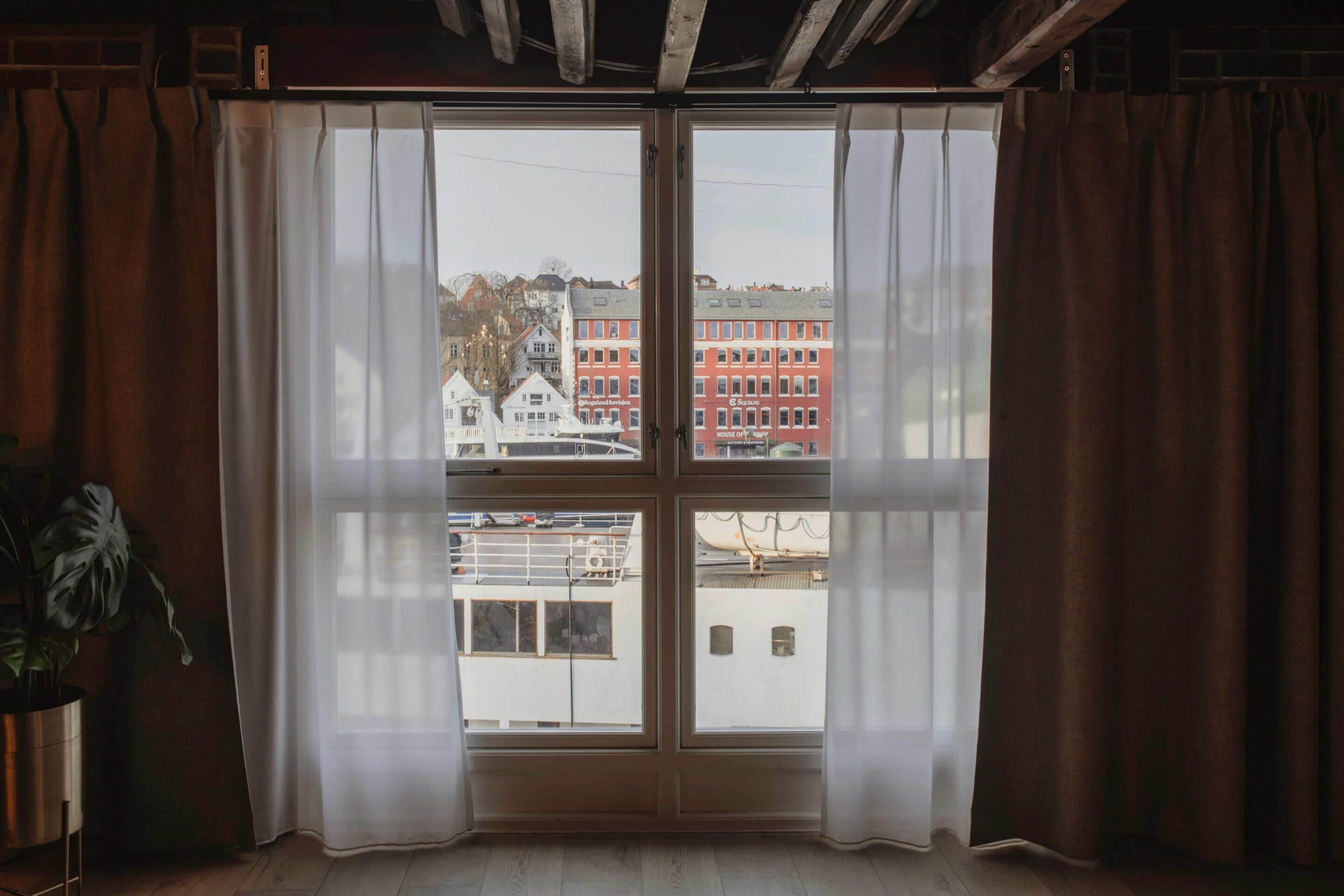 A room with a large window, curtains, a potted plant, and a view of buildings.