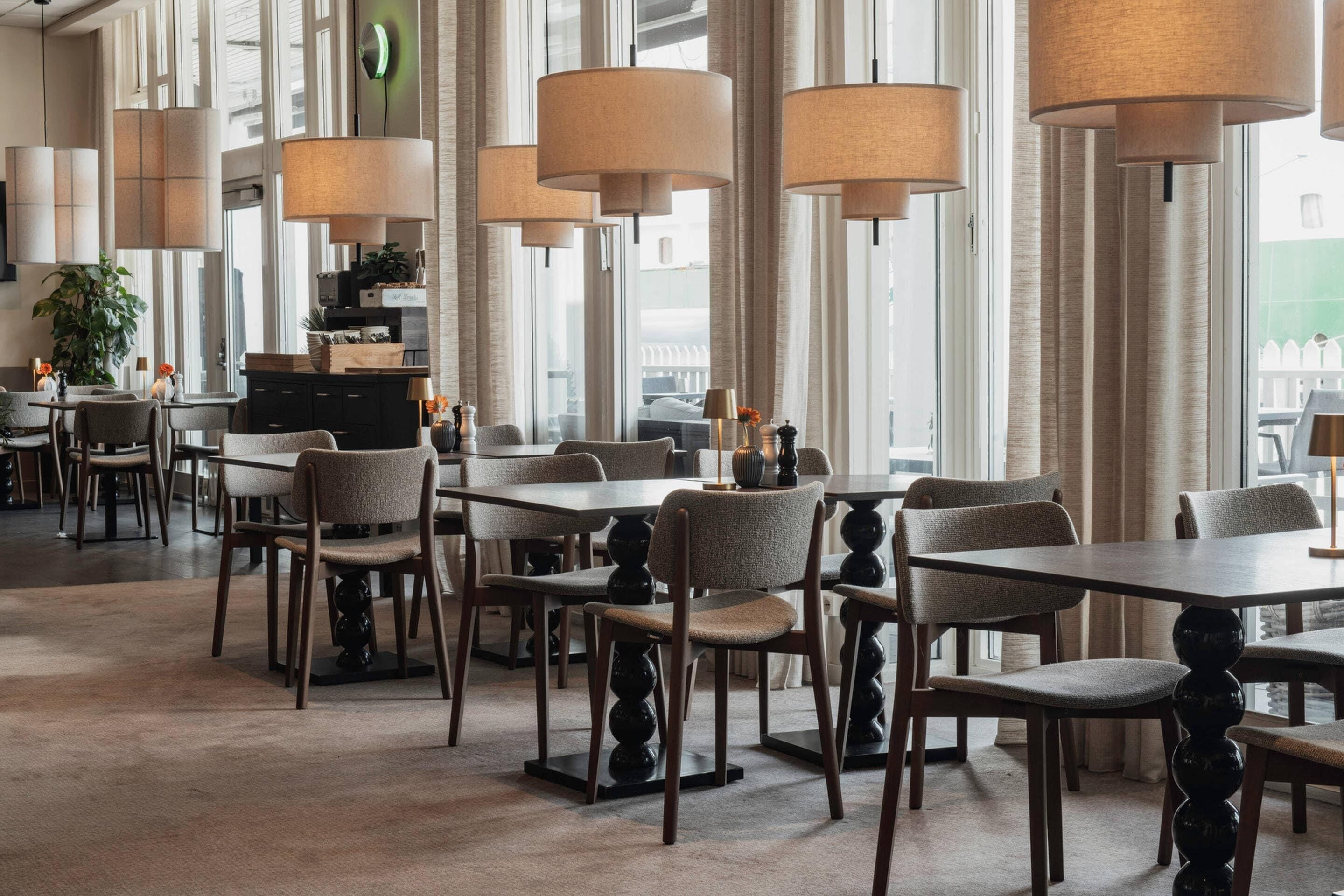 Un'area ristorazione di un hotel con diversi tavoli e sedie, ampie finestre e lampadari.