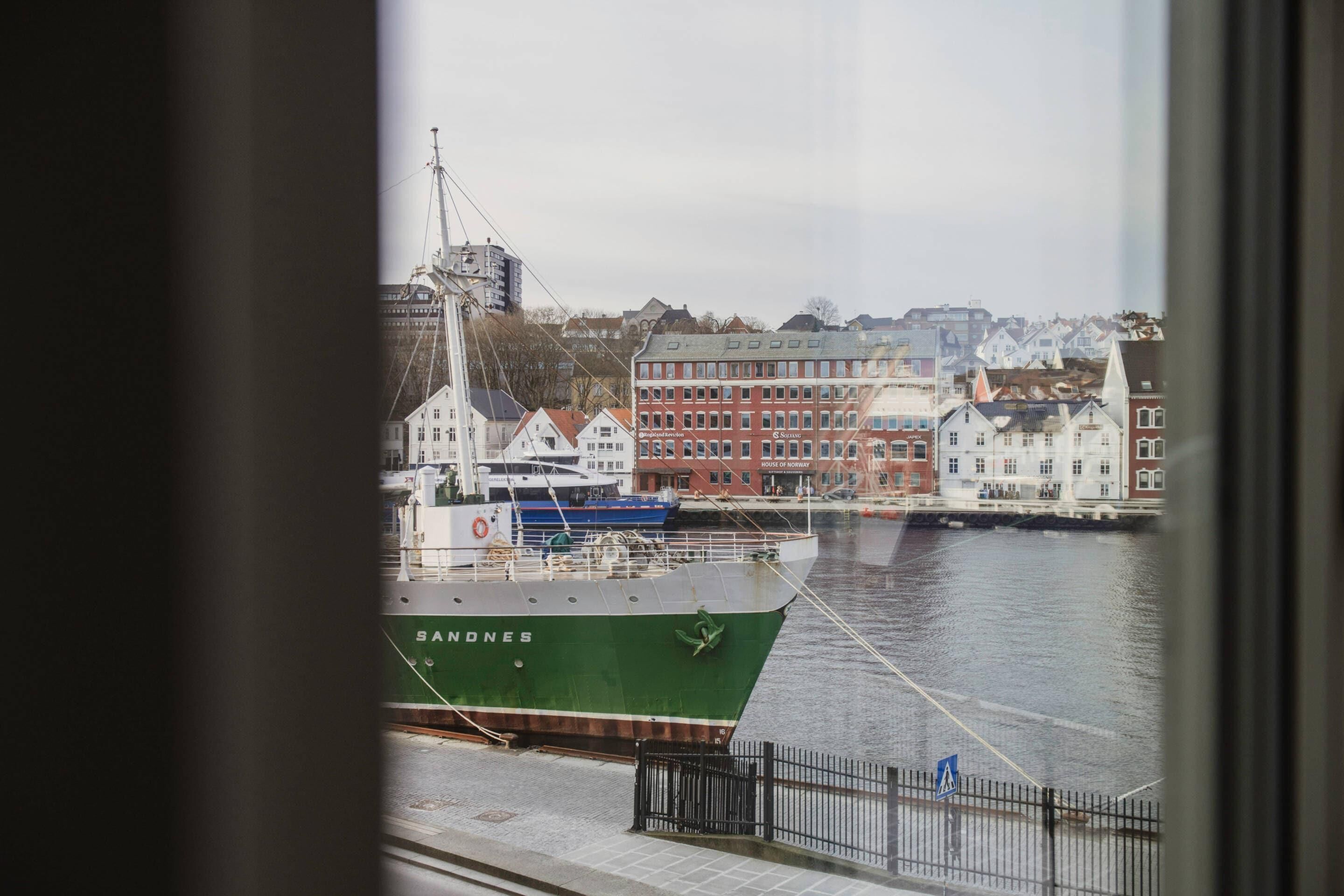 A harbor with a green ship named 'SANDNES' docked, buildings in the background, and a view through a window.