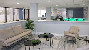 Eine moderne Lobby mit einer Couch, Sesseln und einem Couchtisch. An der Rezeption leuchtet ein grünes Licht, und in der Ecke steht eine Pflanze.