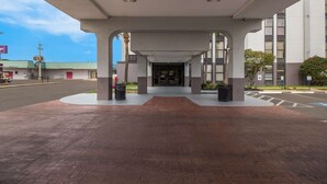 A hotel entrance with a covered walkway, a parking lot, and a building with a green roof in the background.