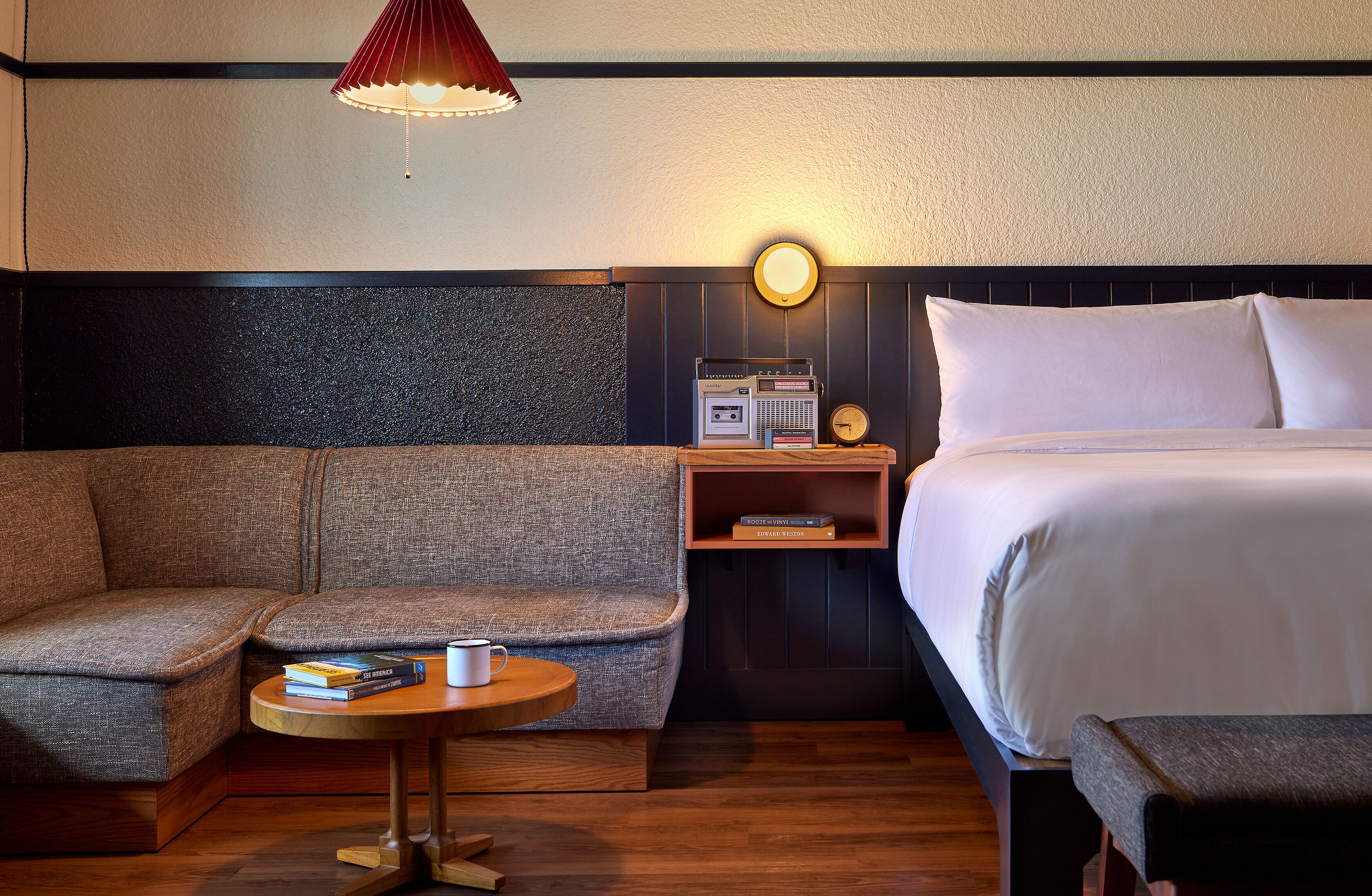 Habitación de hotel con sofá, una mesita redonda con un libro y una taza, una mesita de noche con una radio, y una cama con ropa blanca.