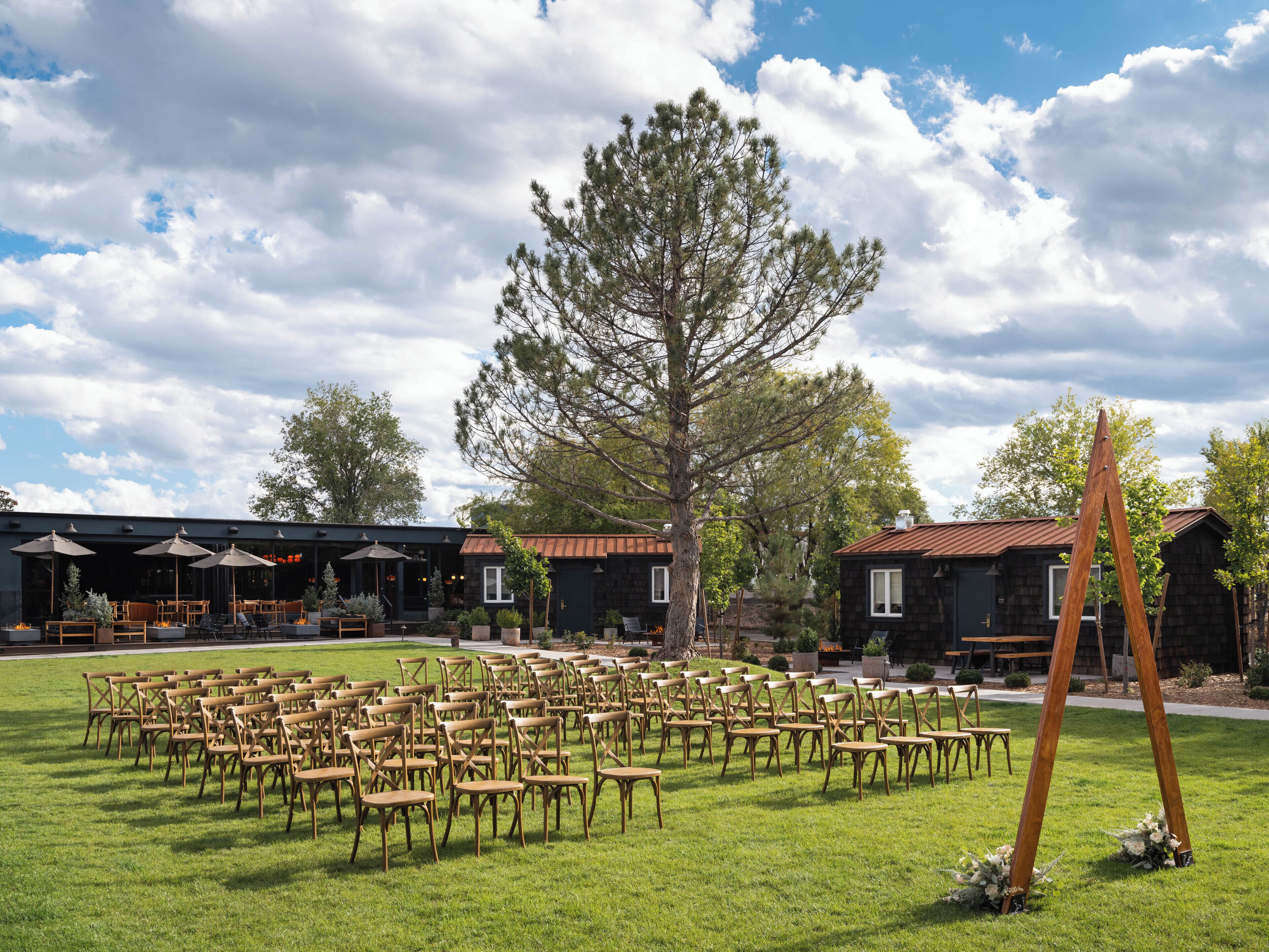 Un espace extérieur aménagé avec des rangées de chaises et de tables en bois, un grand arbre et, au fond, un bâtiment doté d’un toit en métal.