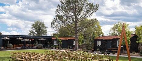 Un espace extérieur aménagé avec des rangées de chaises et de tables en bois, un grand arbre et, au fond, un bâtiment doté d’un toit en métal.