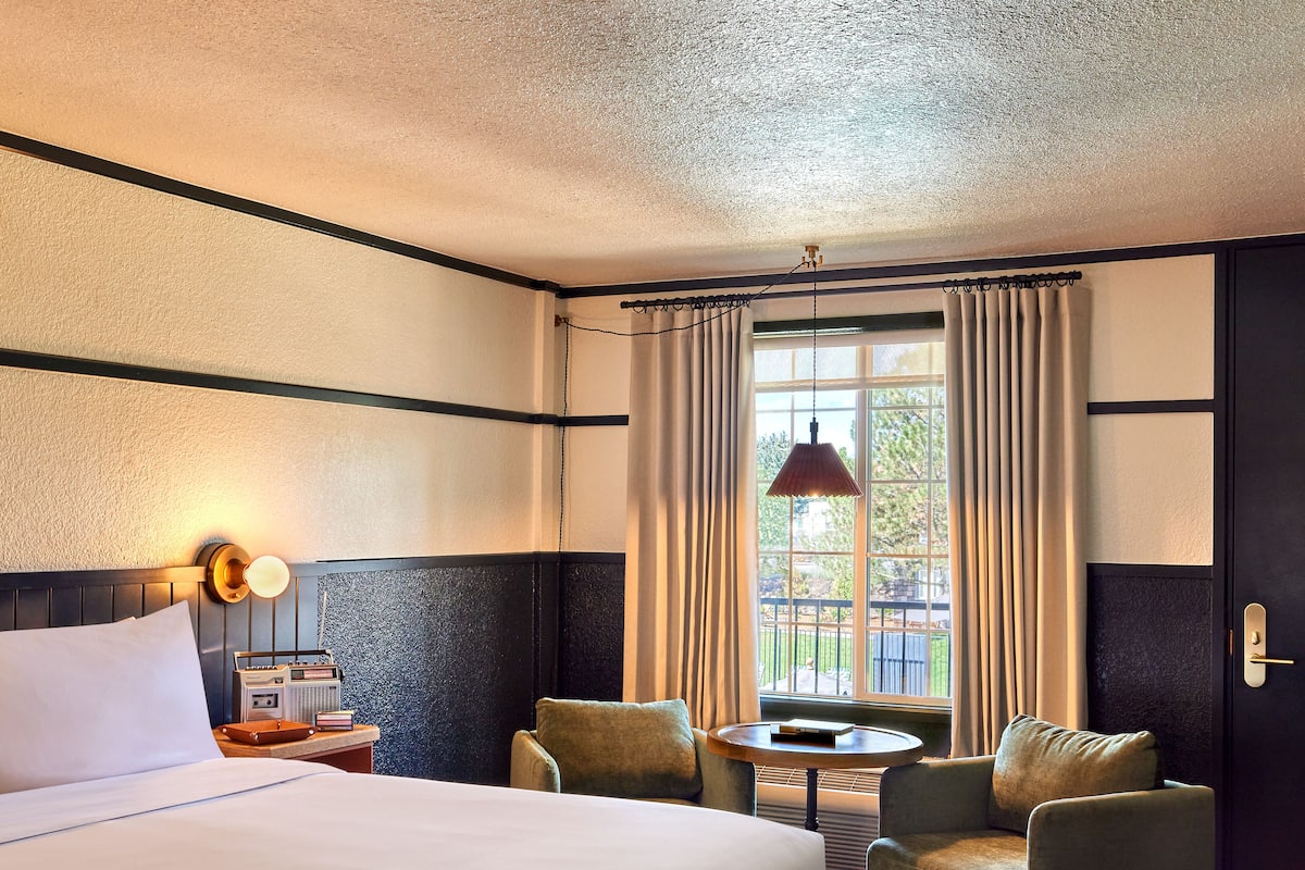 A hotel room with a large bed, two armchairs, a small table, a window with curtains, and a wall-mounted lamp.