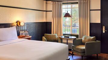 A hotel room with a large bed, two armchairs, a small table, a window with curtains, and a wall-mounted lamp.