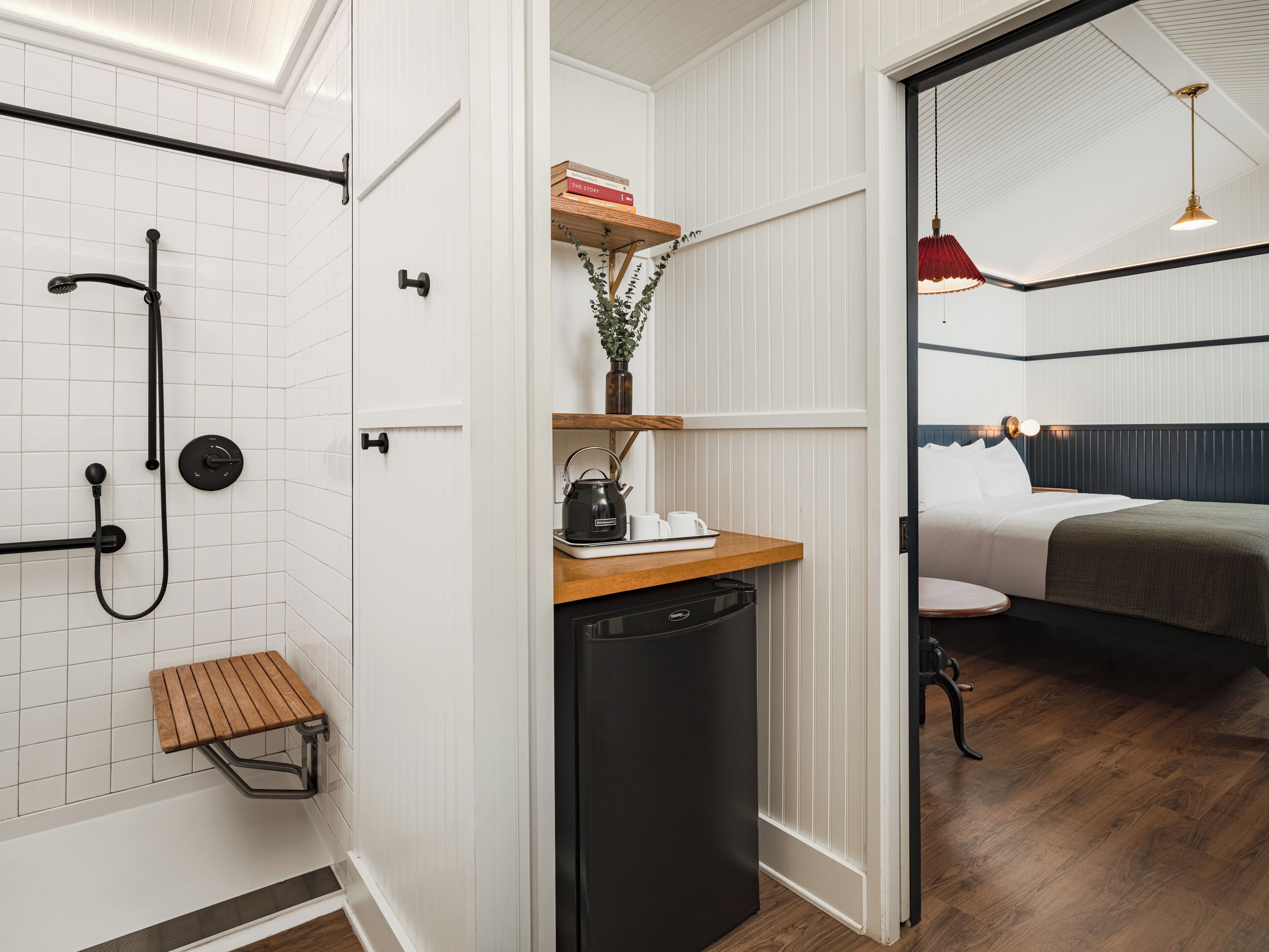 A compact hotel room with a shower, a small wooden stool, a compact kitchen area with a kettle, and a bed with a dark headboard.