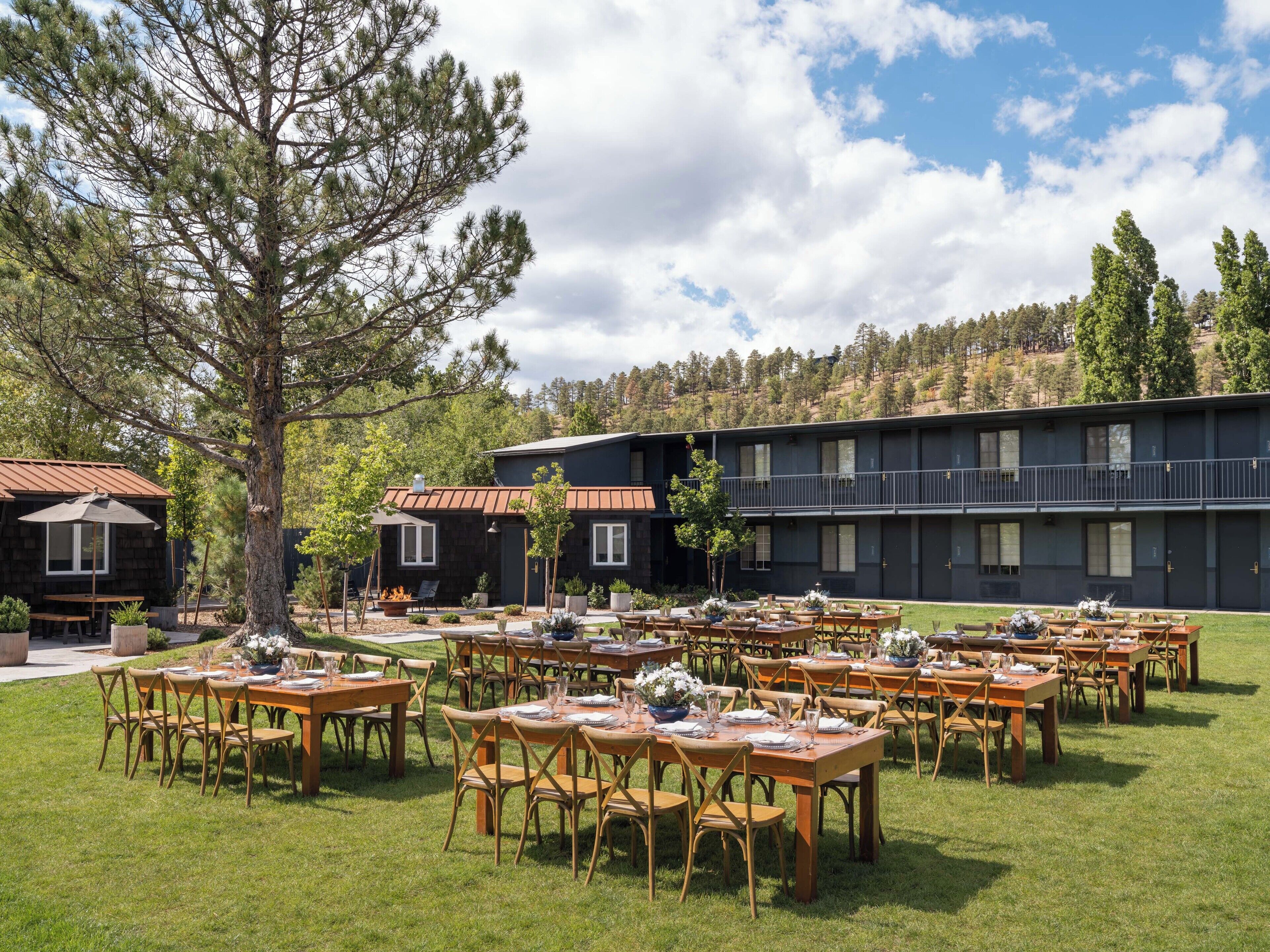 An outdoor dining area with multiple tables and chairs, surrounded by trees and a building with balconies.