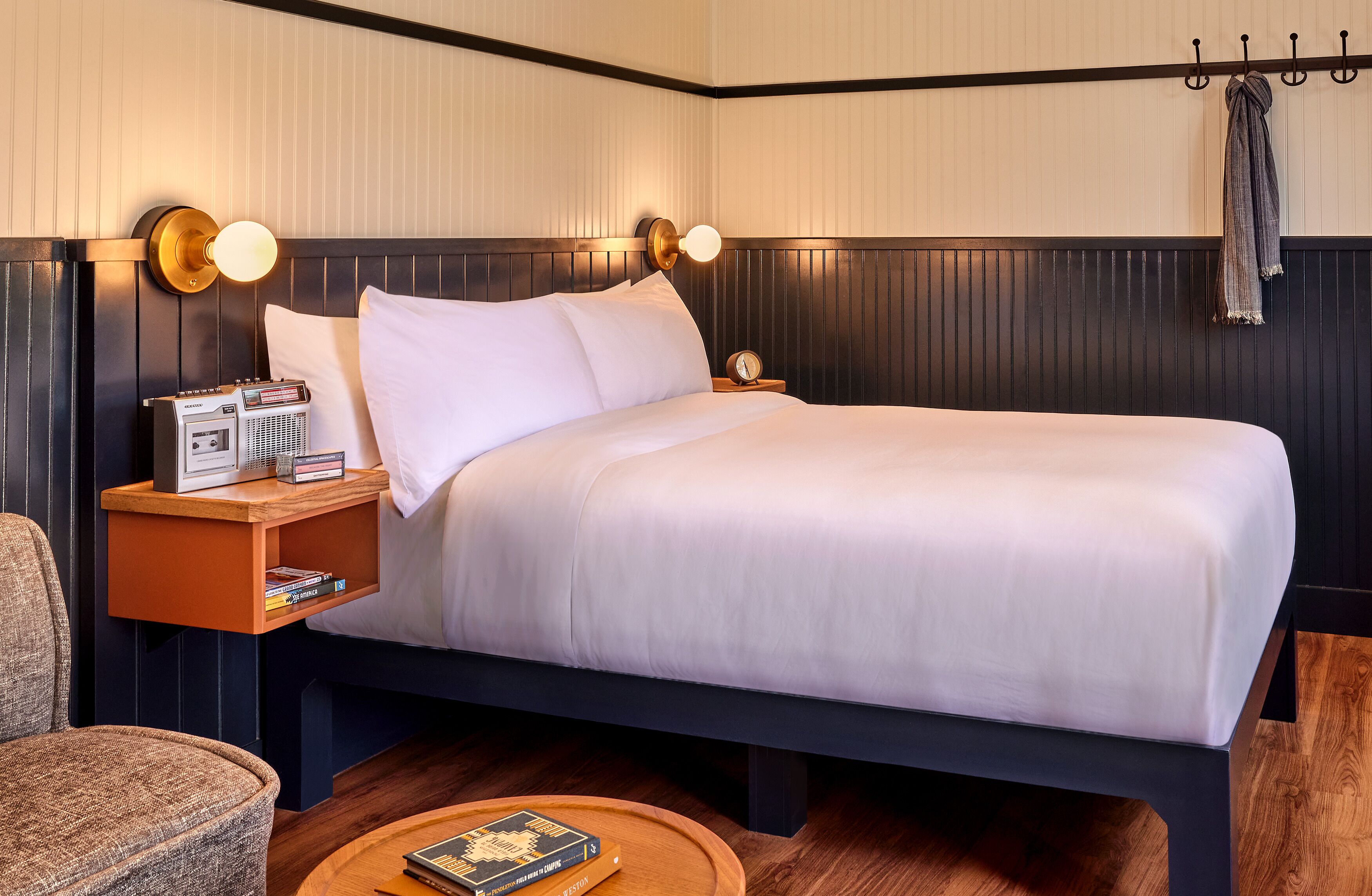 A neatly made bed with white linens, a bedside table with a radio and books, a chair, and a wooden floor.