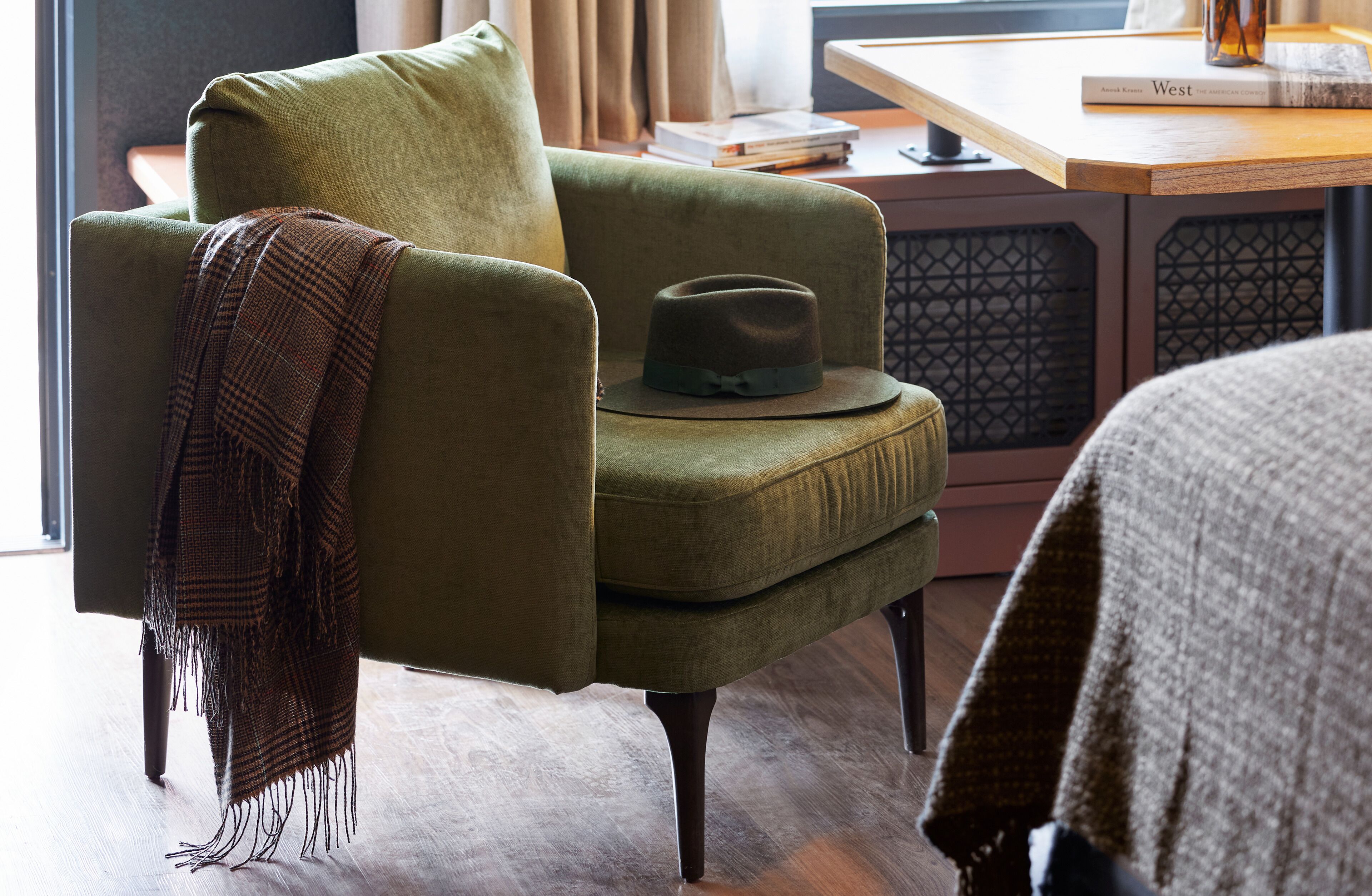 Un sillón verde con un sombrero, una manta y un libro sobre una mesa de madera.