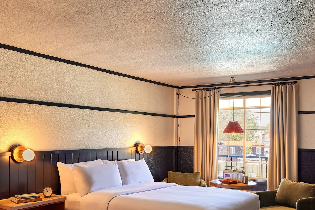 A hotel room with two beds, a nightstand with a lamp, a chair, a window with curtains, and a view of the outside.