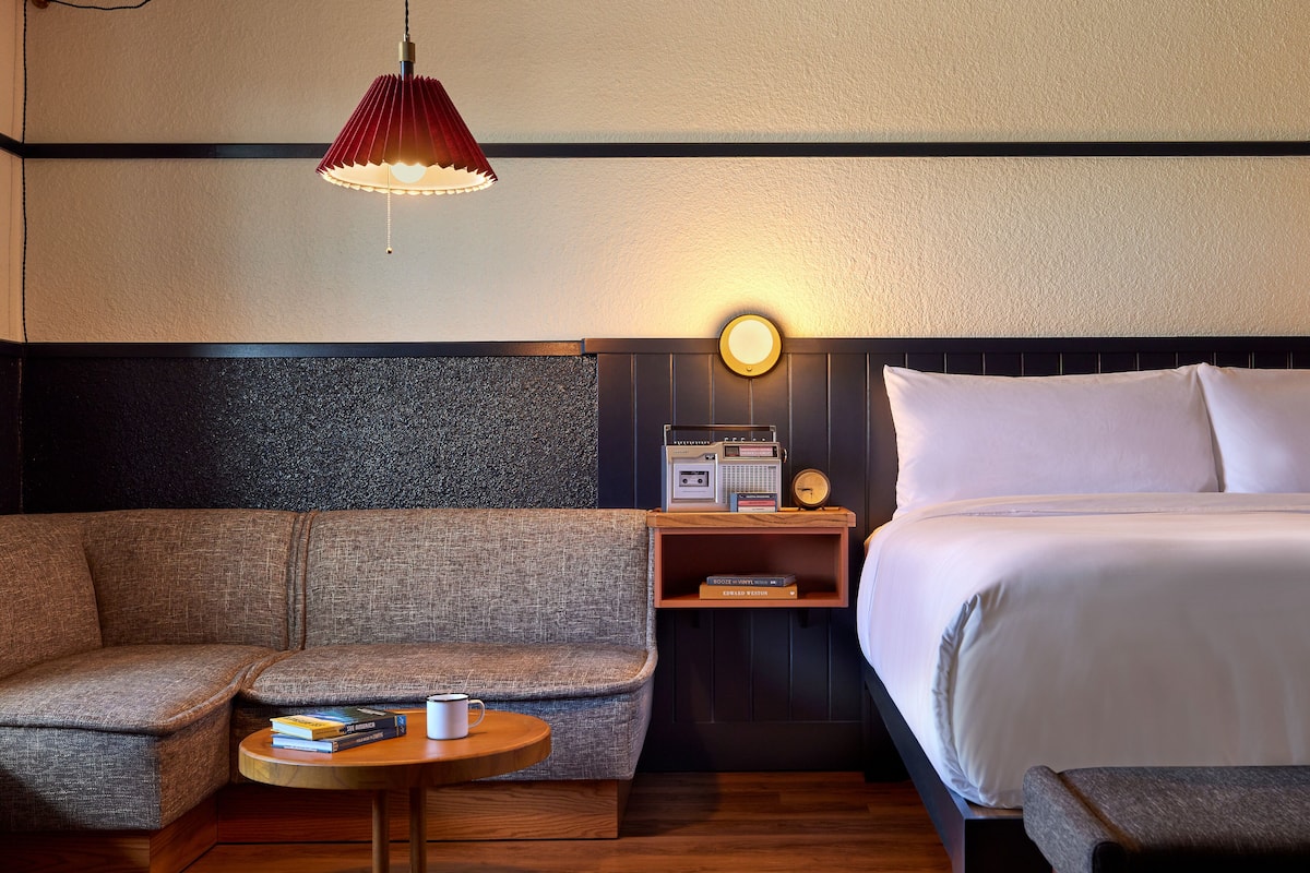 A hotel room with a sofa, a small round table with a book and a mug, a bed with white linens, a nightstand with a radio and clock, and a red pendant light.