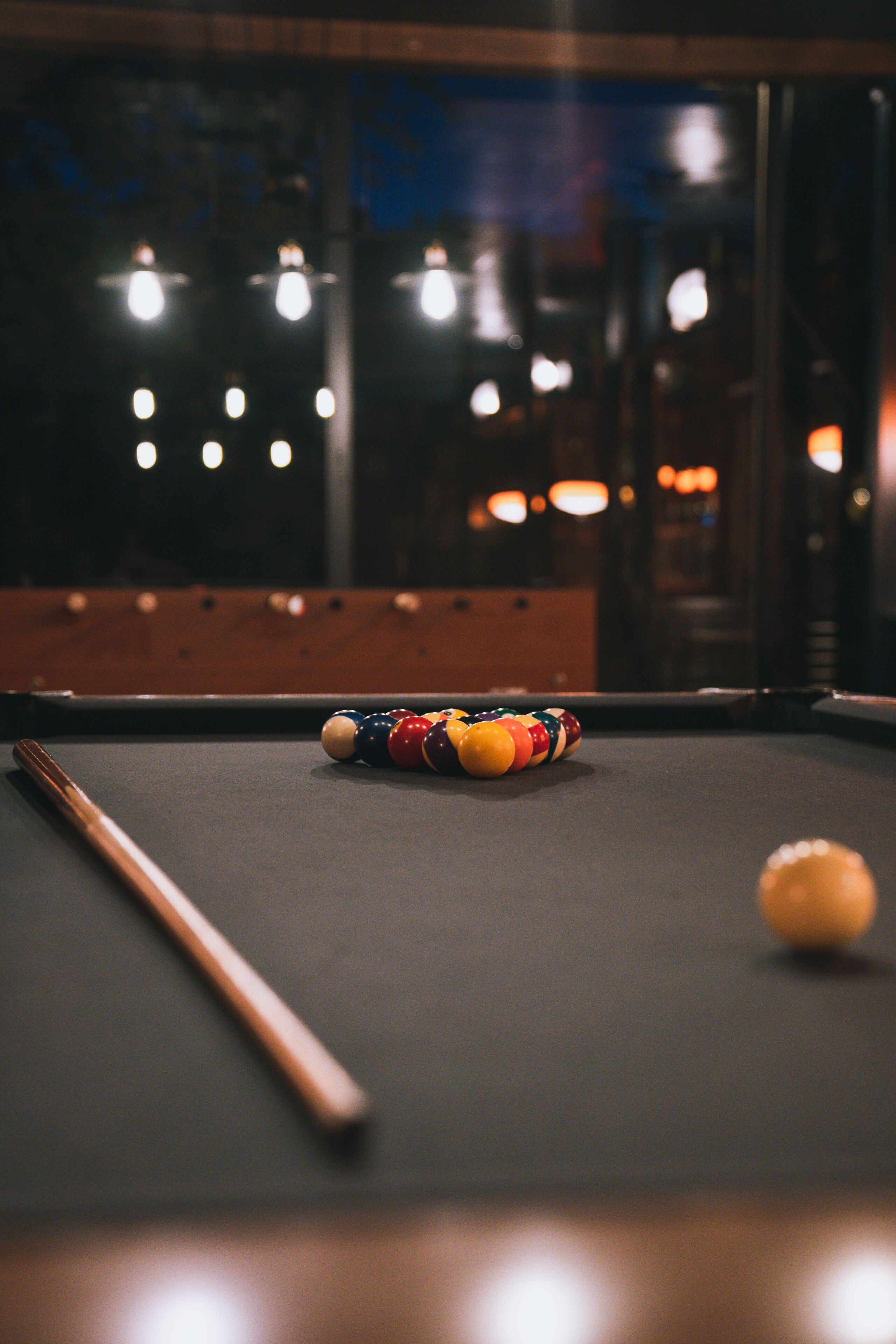 A pool table with a cue stick and balls, a window with lights, and a wooden table edge.