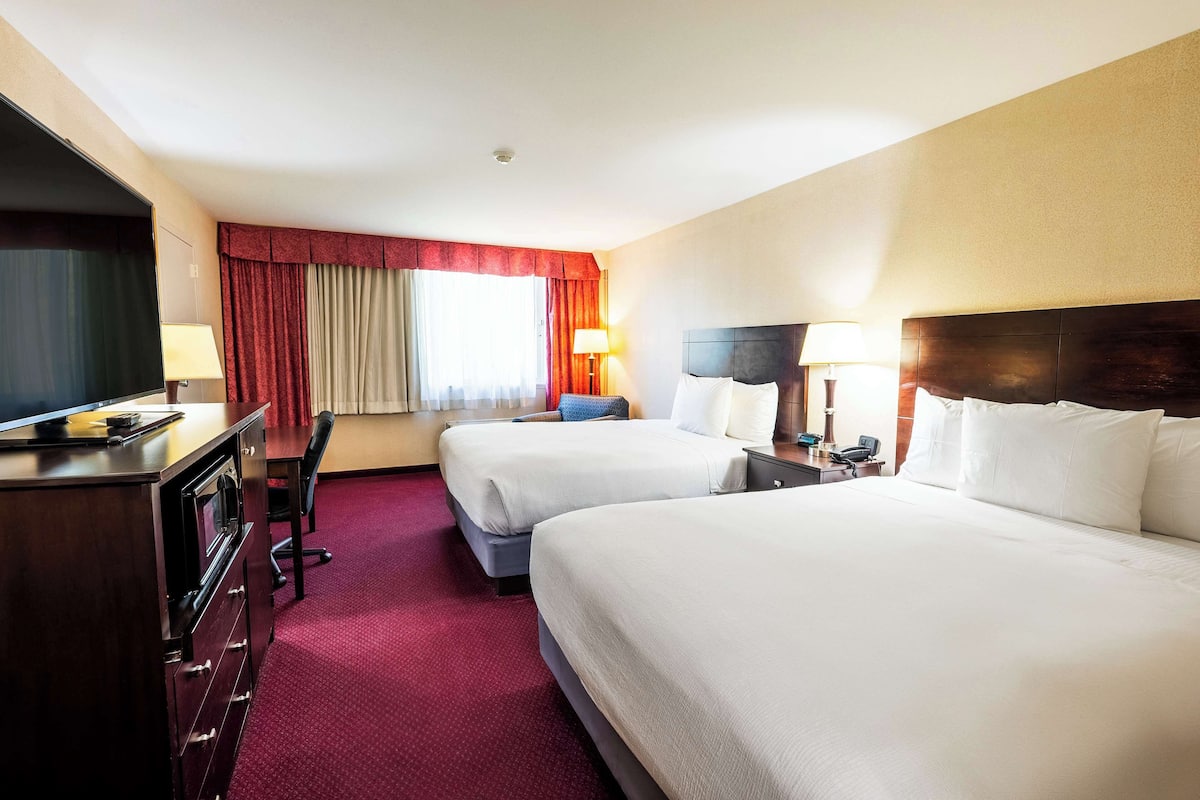 Quarto de hotel com duas camas, uma escrivaninha com televisão, uma luminária e uma janela com cortinas.