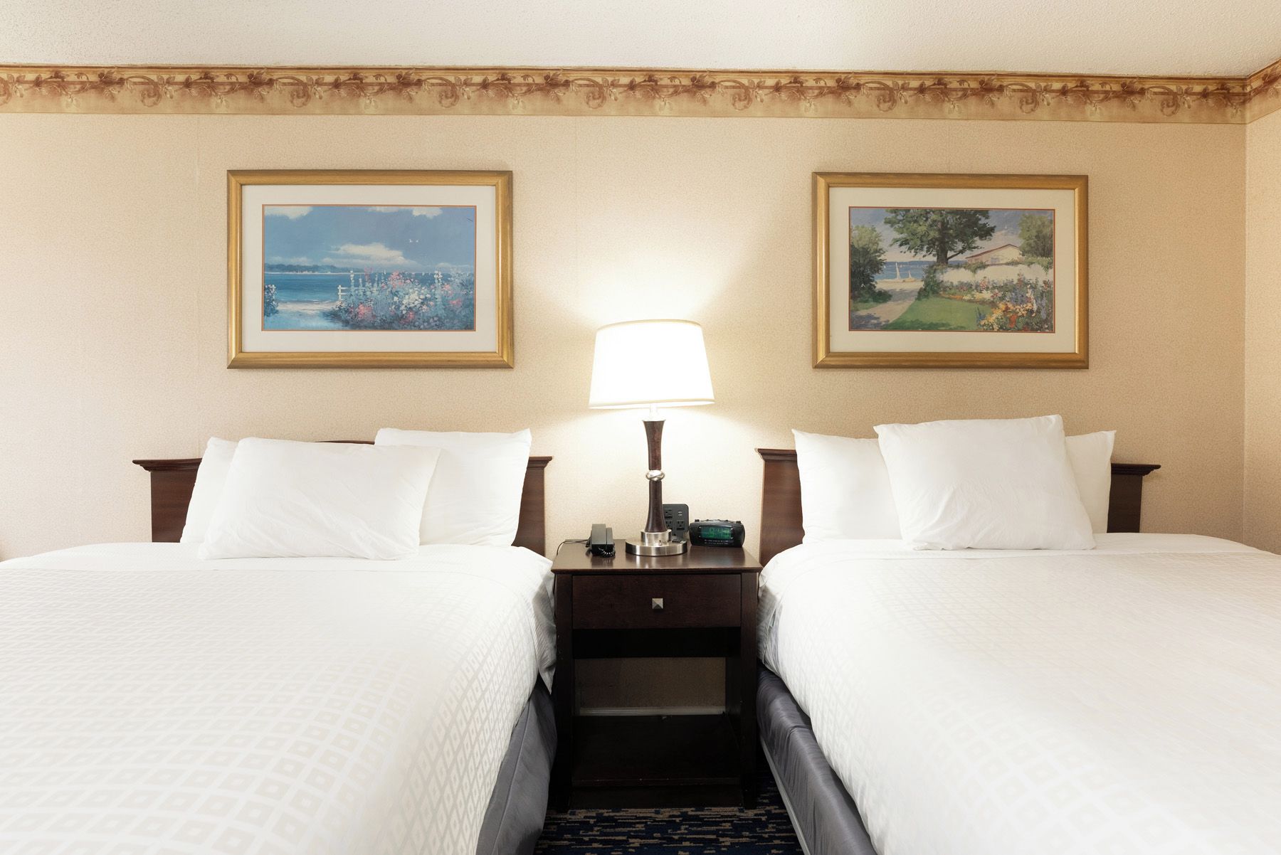 A hotel room with two beds, a nightstand with a lamp, and two framed pictures on the wall.