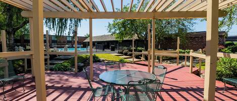Un área de patio techada con una mesa de vidrio y sillas de metal, rodeada por una pérgola de madera.