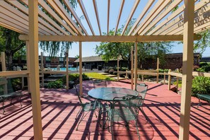 A covered patio area with a glass table and metal chairs, surrounded by a wooden pergola.