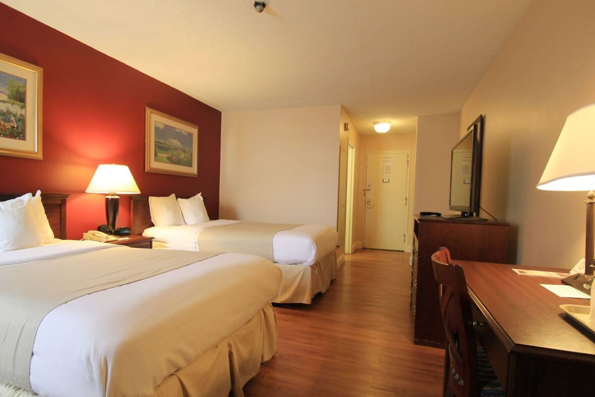 A hotel room with two beds, a desk, a chair, a lamp, and a painting on the wall.