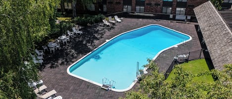 A rectangular swimming pool surrounded by lounge chairs and tables, with a brick building in the background.