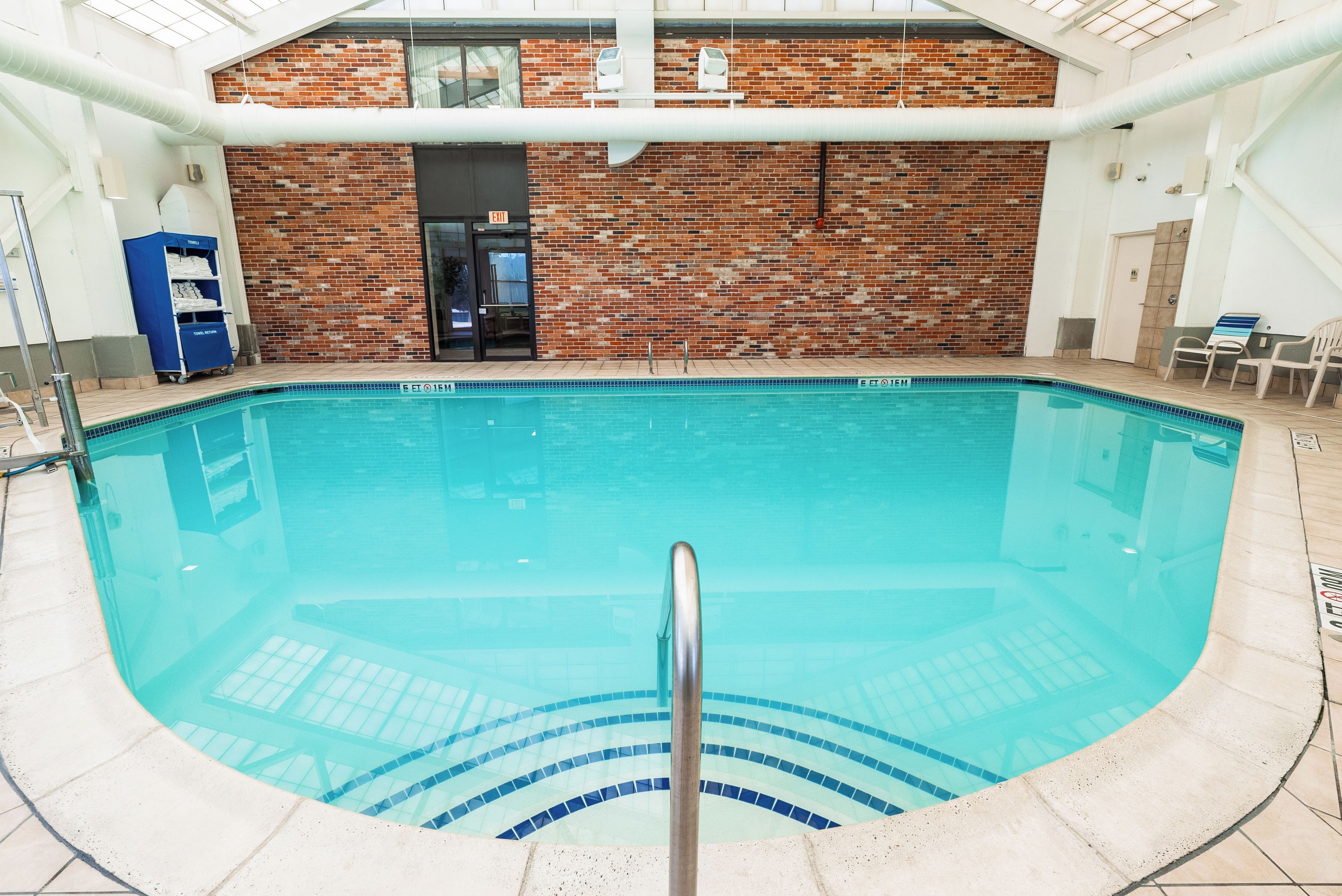 An indoor swimming pool with a brick wall backdrop, a clear pool edge, and a visible exit sign.