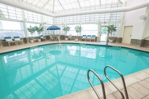 An indoor swimming pool with clear glass walls, lounge chairs, and potted plants.