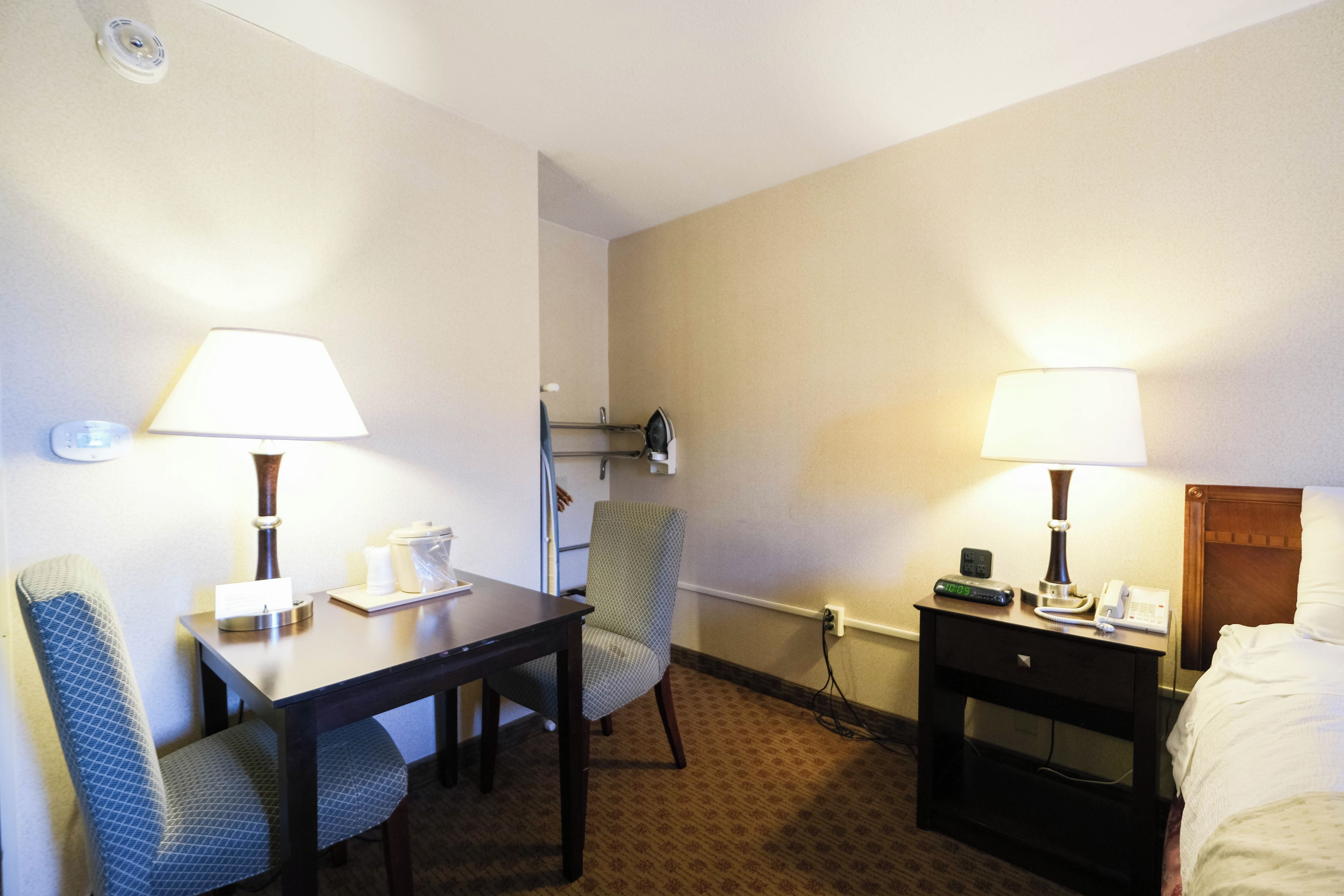A hotel room with a bed, two chairs, a desk, a nightstand, a lamp, a clock, and a wall-mounted heater.