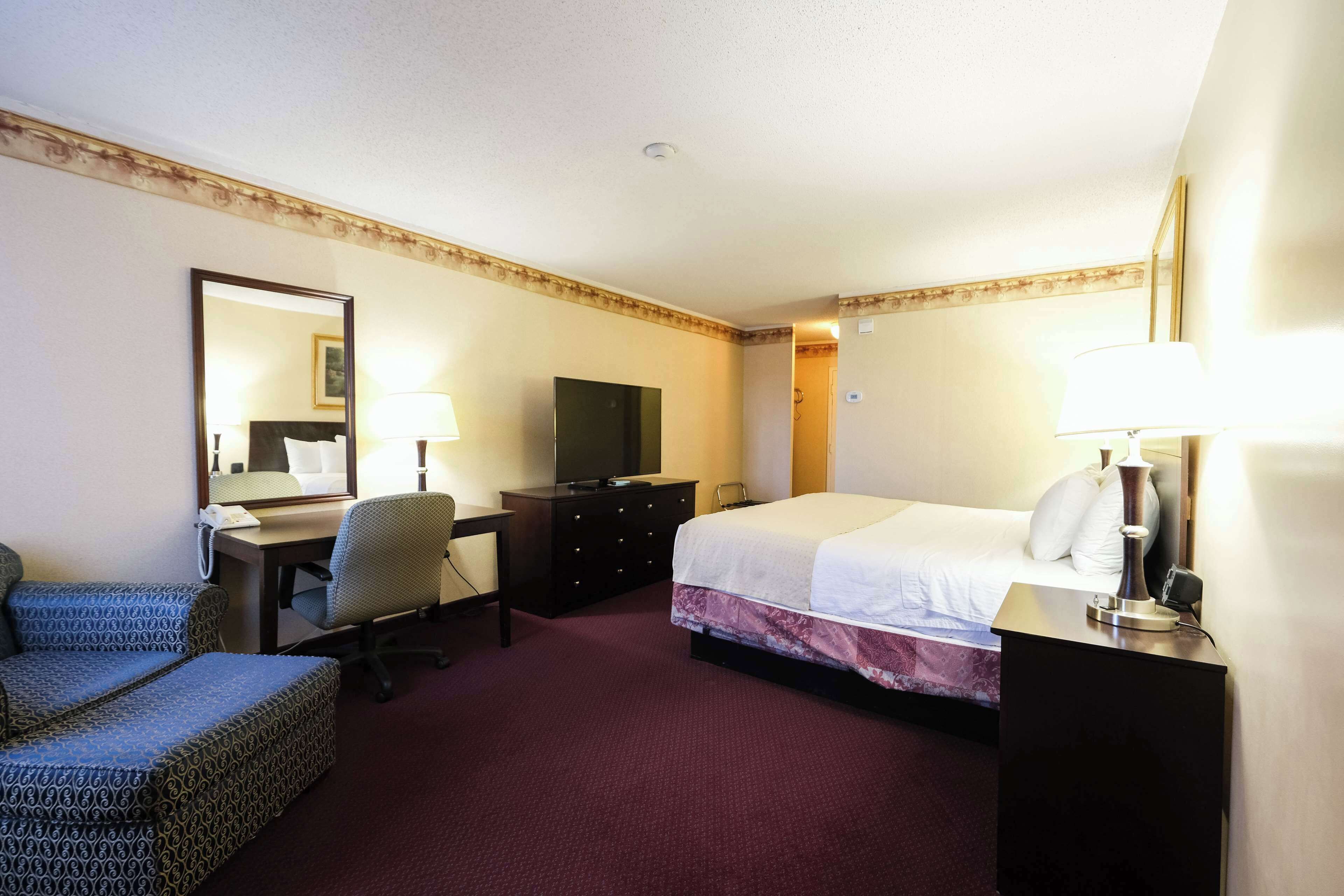 Habitación de hotel con cama, escritorio, silla, televisor, cómoda y espejo.