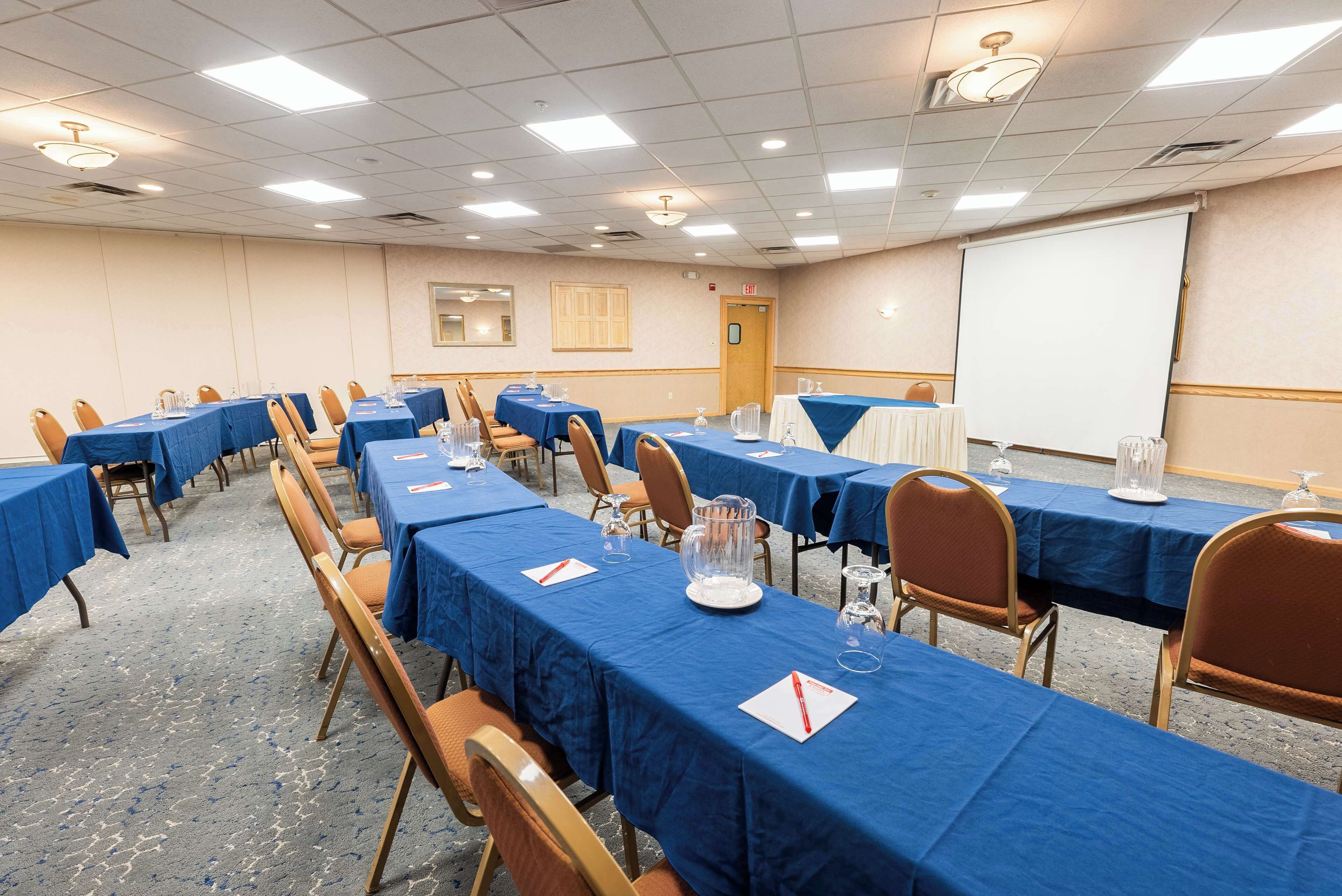 Ein Konferenzraum mit mehreren Tischen und Stühlen, ausgestattet mit Wasserkännchen und Gläsern sowie einer Projektionsleinwand.