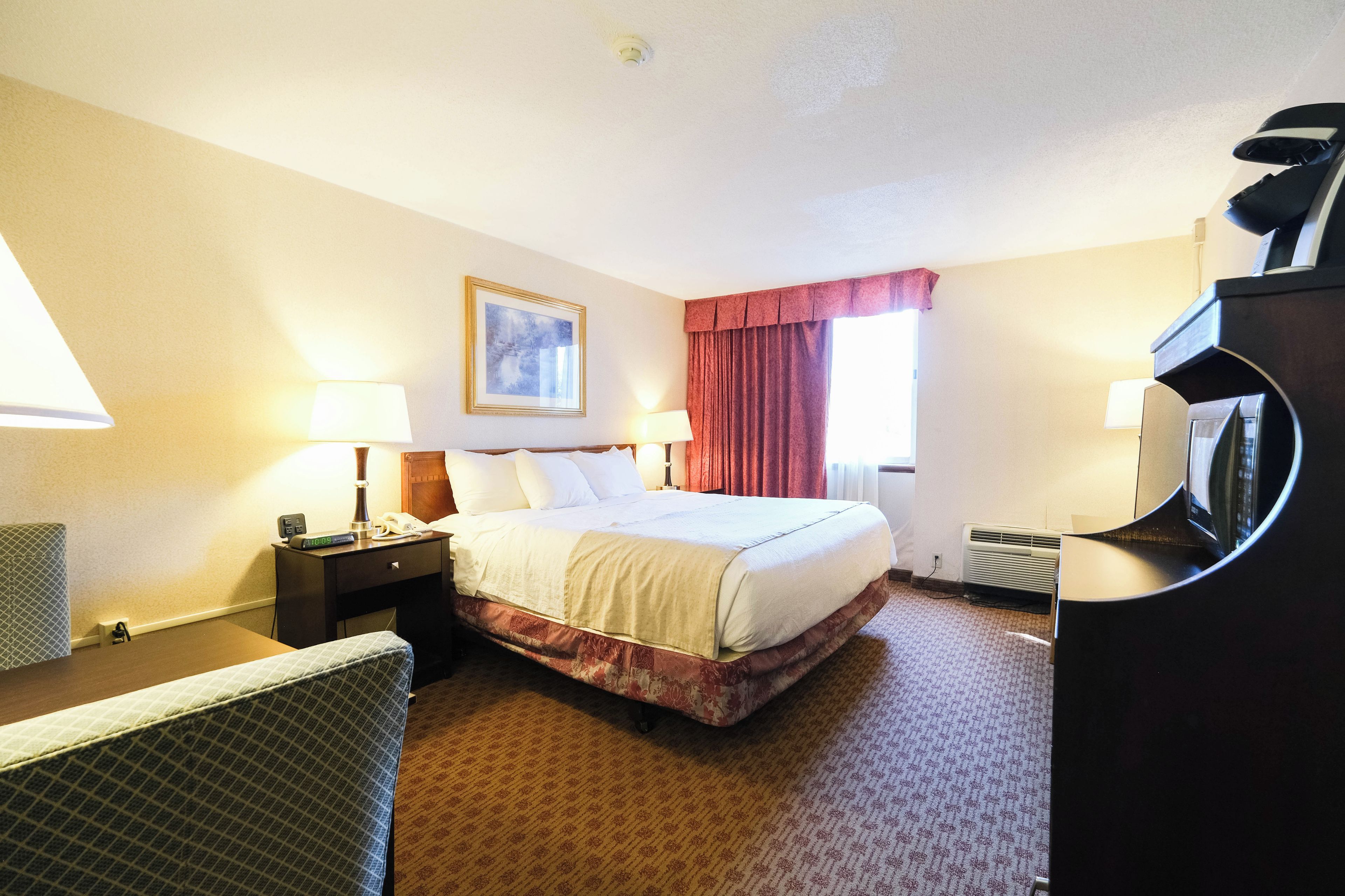 A hotel room with a bed, two bedside tables, a desk, a chair, a window with curtains, a wall clock, and a painting.