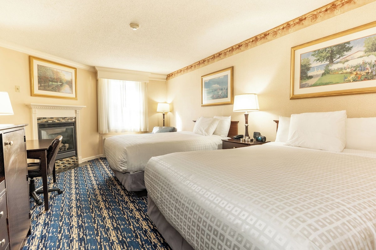 A hotel room with two beds, a desk, a chair, a fireplace, and framed artwork on the walls.