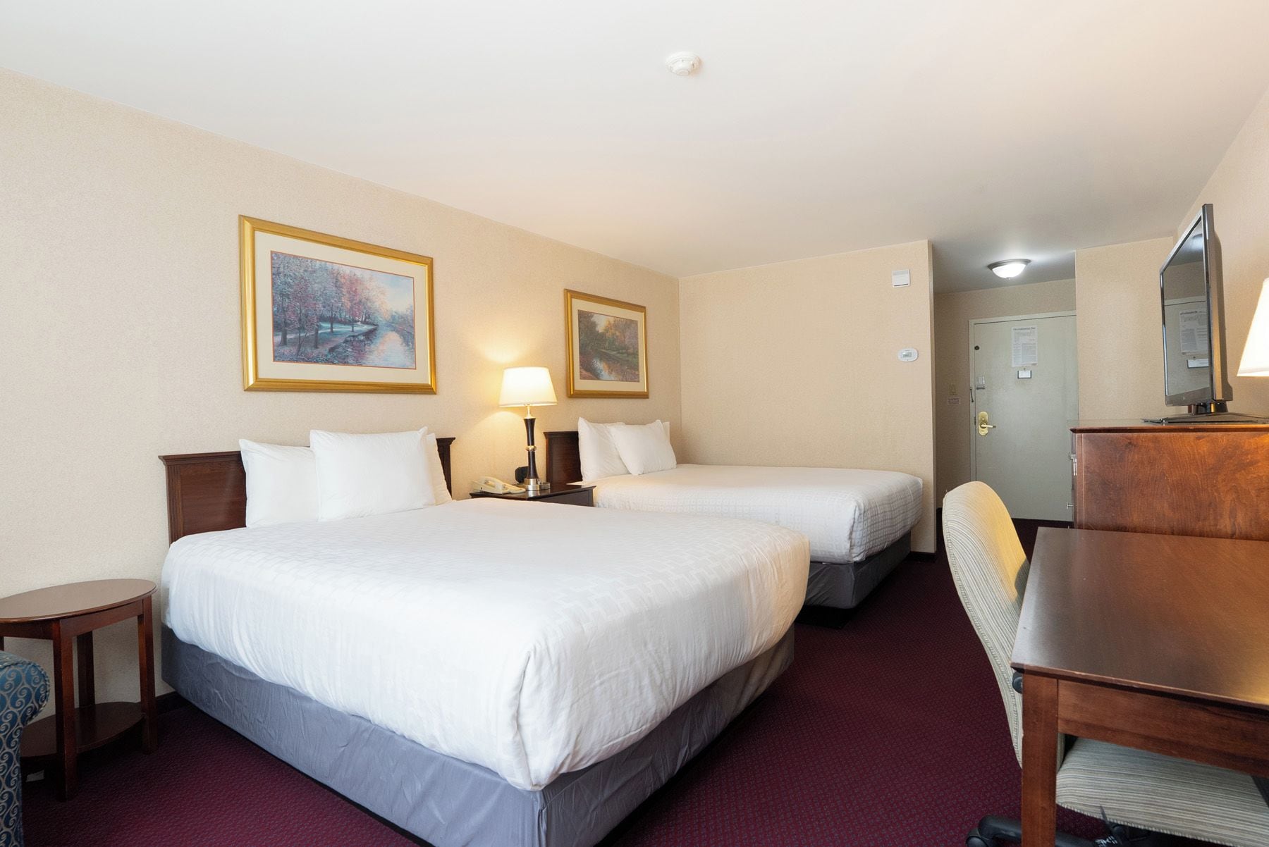 A hotel room with two beds, a desk, a chair, a lamp, and two paintings on the wall.