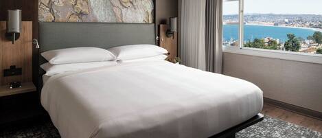A large bed with white linens, a stone wall art piece above it, a nightstand with a lamp, and a view of the sea through a window.