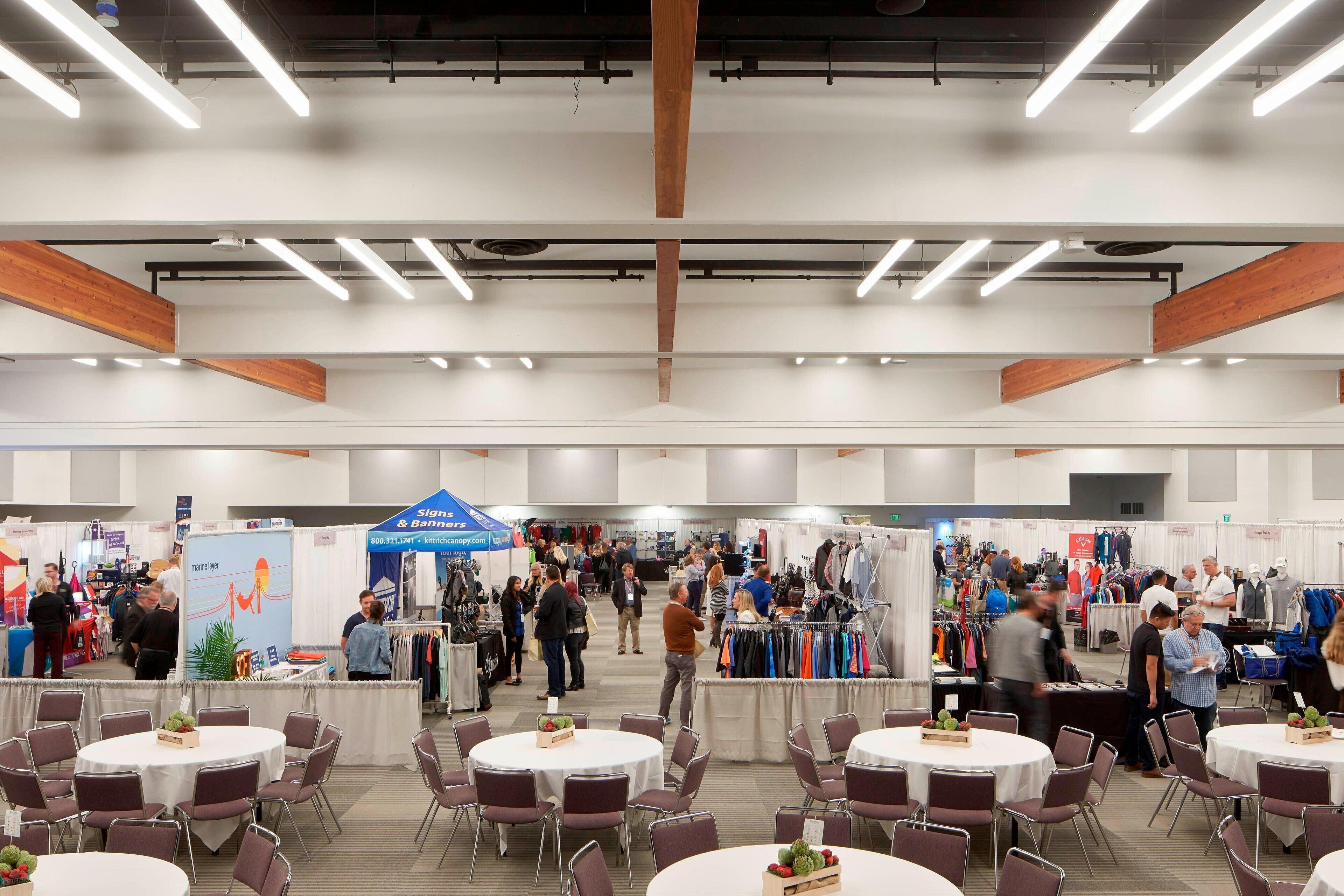 Un vaste espace intérieur aménagé pour des événements, avec des tables et des chaises rondes, des poutres en bois au plafond, et des participants qui déambulent parmi les stands.