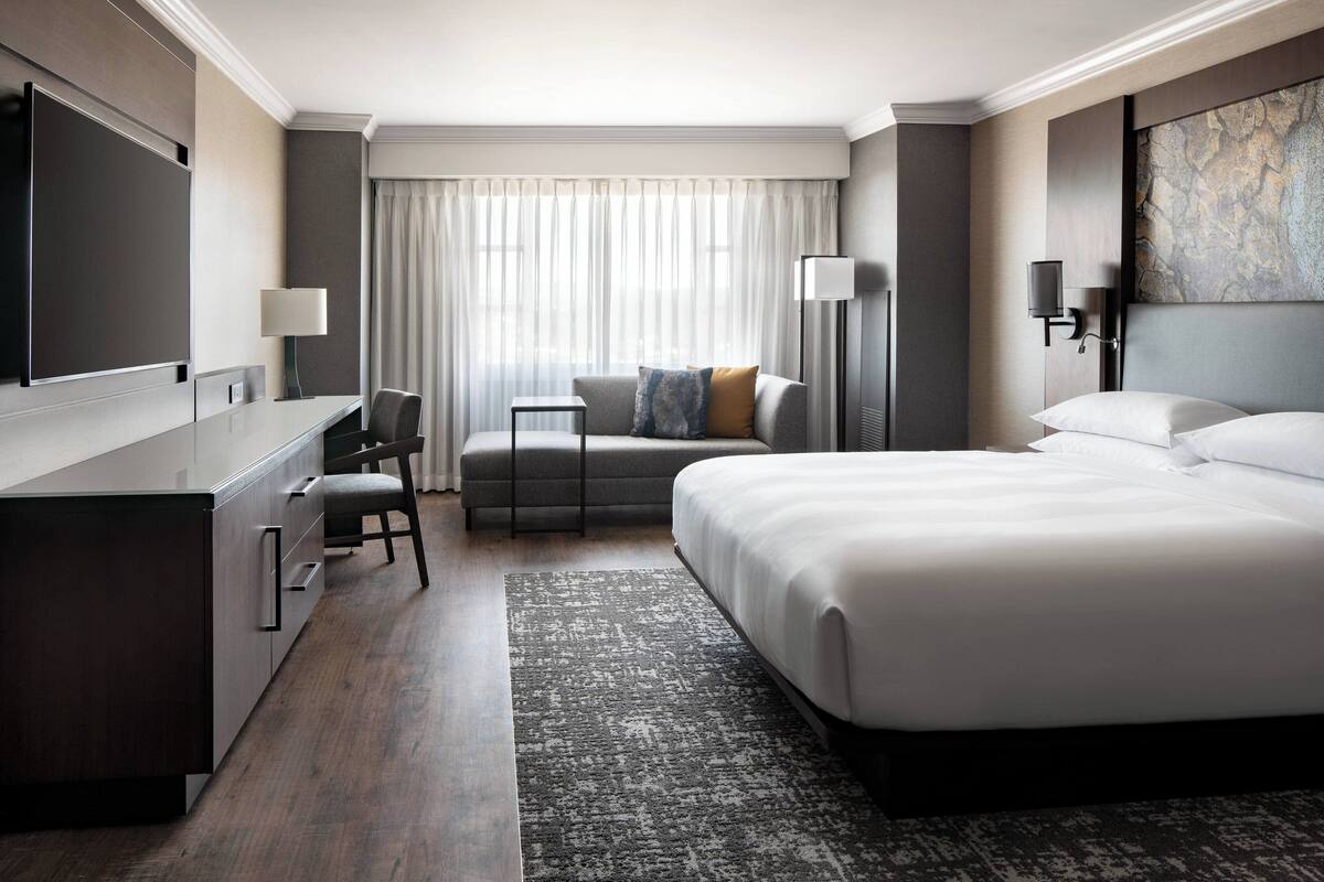 Una habitación de hotel moderna con una cama grande, un escritorio, una silla, una mesita pequeña y un amplio ventanal con cortinas.