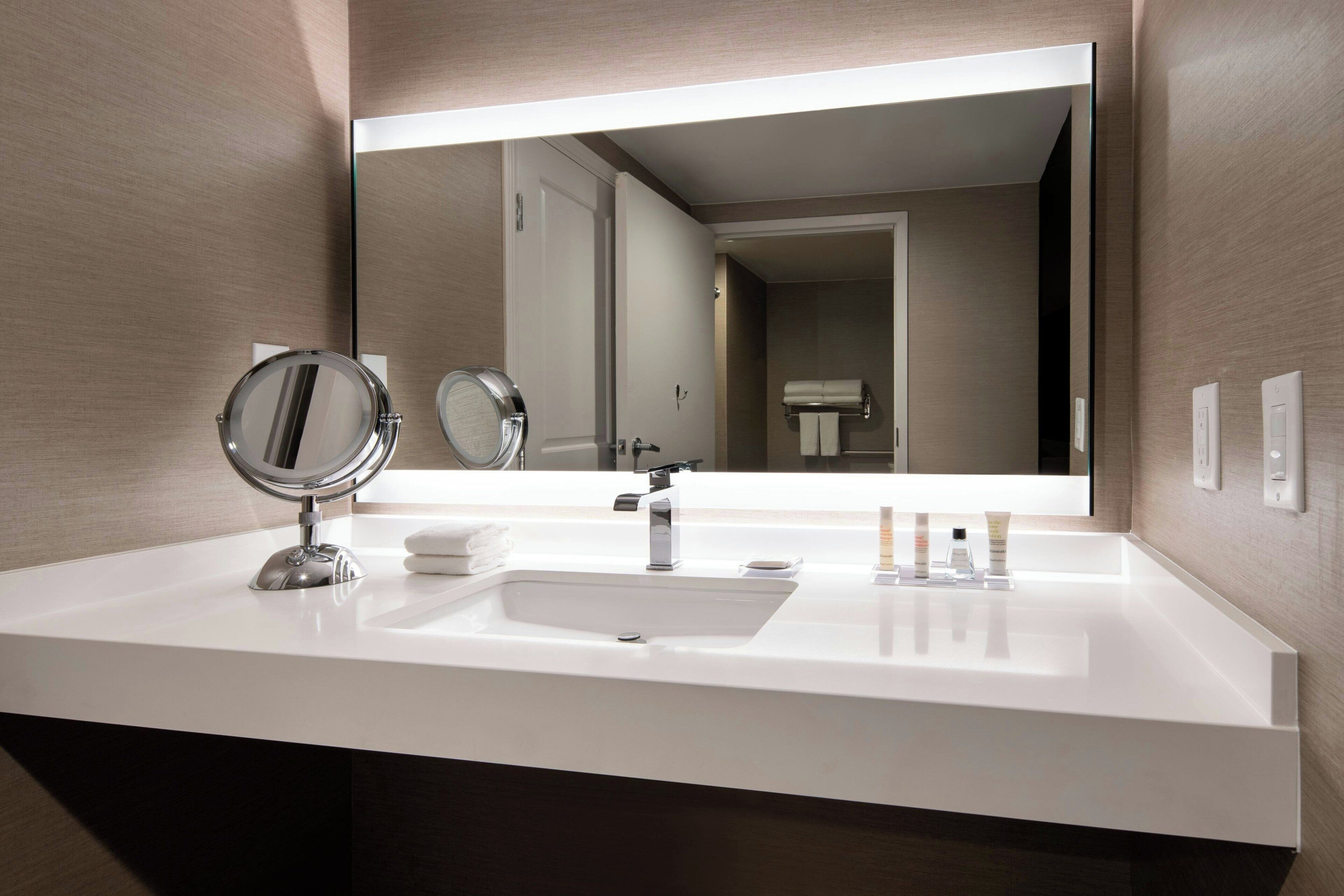 Un baño moderno con un lavamanos blanco, dos espejos, un perchero para toallas y artículos de aseo.